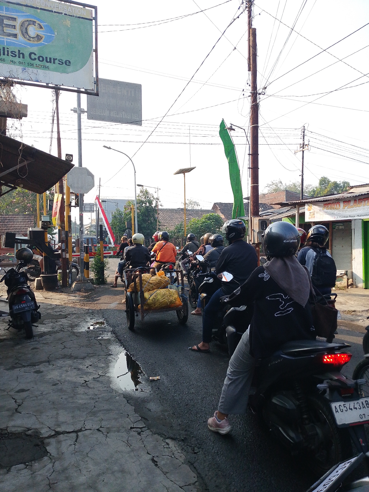 The image captures a street scene in Kediri City, Indonesia, likely near a railway crossing. In the foreground, a person on a black motorcycle with the license plate AG5443AB is visible from the side. The motorcycle's taillight is illuminated. To the left, another black motorcycle, license plate AC 4580 ECR, is parked. Further back, a small cart filled with yellow produce is being pulled by a person on a motorcycle. A line of several motorcycles, many with riders wearing helmets, are queuing up. The road surface is asphalt with visible cracks and some puddles of water. In the background, a red and white Indonesian flag is prominently displayed. Utility poles with numerous wires crisscross the sky. Signs are visible, including one for an "English Course" and another indicating "BERHENTI SEBELUM MELINTASI REL," which translates to "STOP BEFORE CROSSING THE RAILWAY." Buildings line the street, some appearing to be shops. The overall impression is a busy everyday scene at a traffic point in a city.