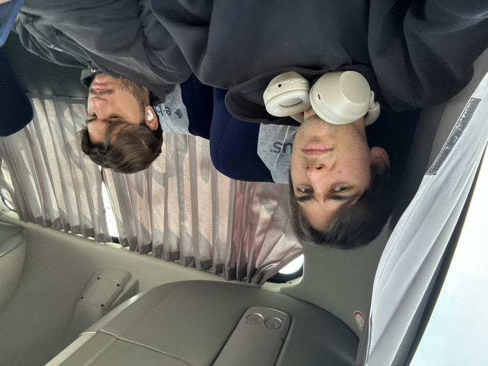 Two young men sit inside a vehicle, possibly a bus or a train. One wears headphones around his neck, while the other has an earbud in. They are seated near a window with closed curtains. The interior of the vehicle includes a gray wall and seating area.