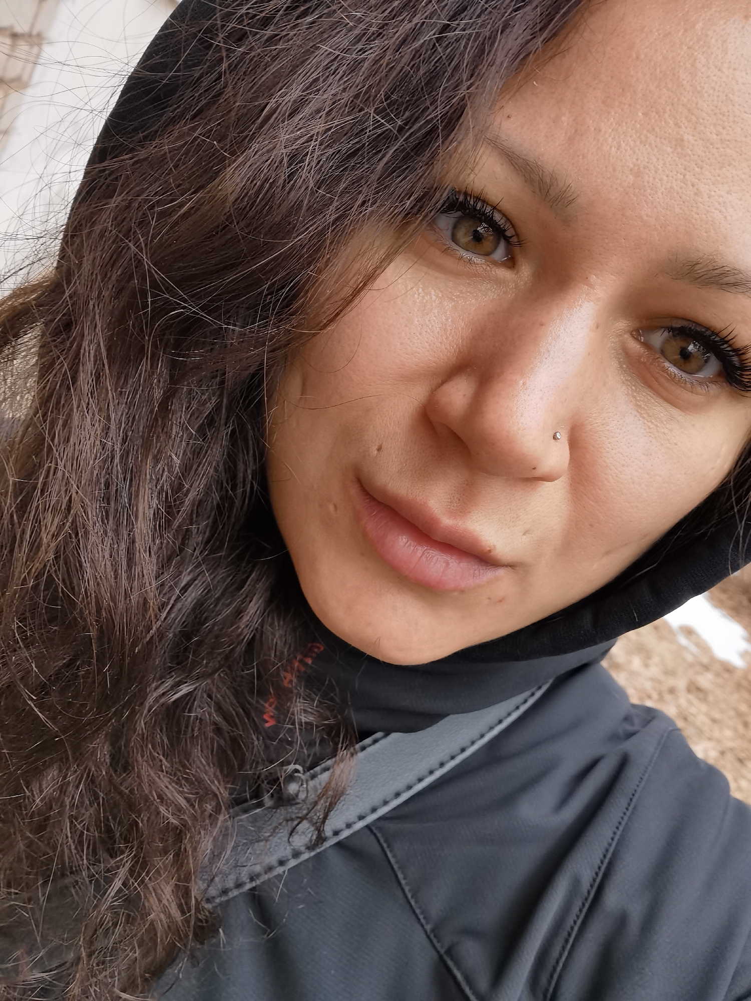 A close-up, selfie-style photograph features a woman with dark brown, wavy hair partially covered by a black hooded jacket. She has fair skin, light brown eyes, and a nose piercing on one side. She is looking at the camera with a slight smile. The background is blurred and shows some indistinct outdoor features, possibly snow or ground cover. The jacket appears to be a dark gray or black athletic or outerwear style with some gray trim. The location is possibly in Chaikovsky, Russia, but there is no conclusive evidence within the image to confirm the exact location.