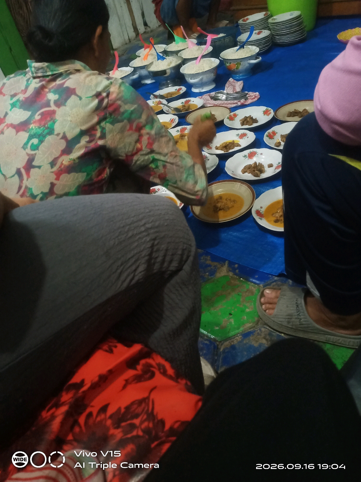 The image depicts a gathering of people, likely in Plosorejo, Indonesia, engaged in a meal served on a blue tarp spread across a floor. Several individuals are partially visible, including a person in a floral shirt, another in grey pants, and someone wearing a pink head covering. The meal is laid out on numerous plates and bowls, featuring what appears to be rice and meat dishes. Utensils are also present. The environment suggests an informal indoor setting, possibly a home or community space, with simple furnishings and lighting.