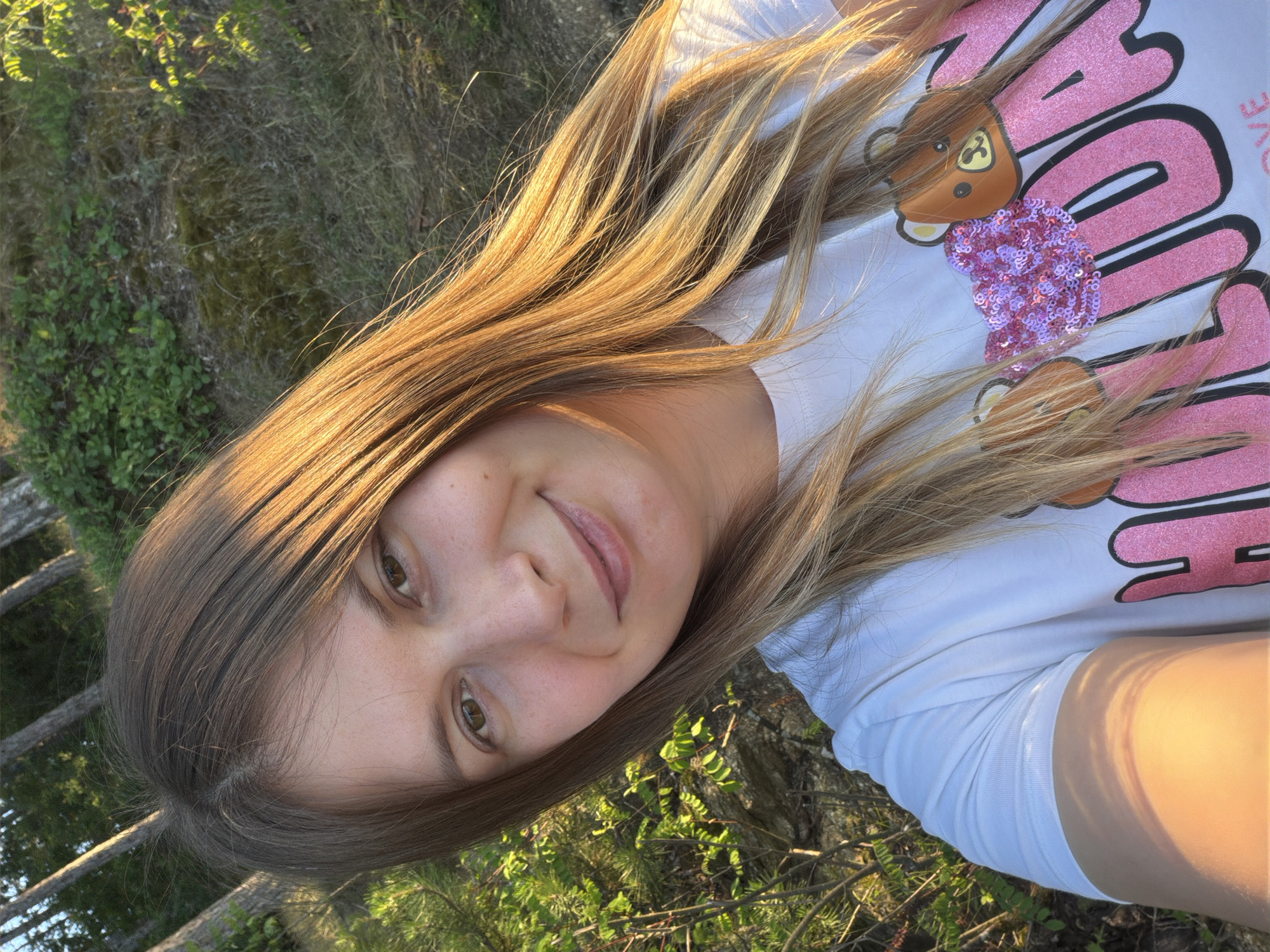 A young woman with long, blonde-brown hair, parted in the middle and falling over her shoulders, smiles directly at the camera. She is wearing a white t-shirt with a graphic design of teddy bears and pink lettering that appears to say "HOLLYDAY". The background suggests an outdoor, natural setting with trees and greenery, consistent with a location like Koprinka, Bulgaria. The lighting indicates it is either early morning or late afternoon, with warm sunlight illuminating the woman's face and hair. The overall impression is a candid, outdoor portrait taken in natural light.
