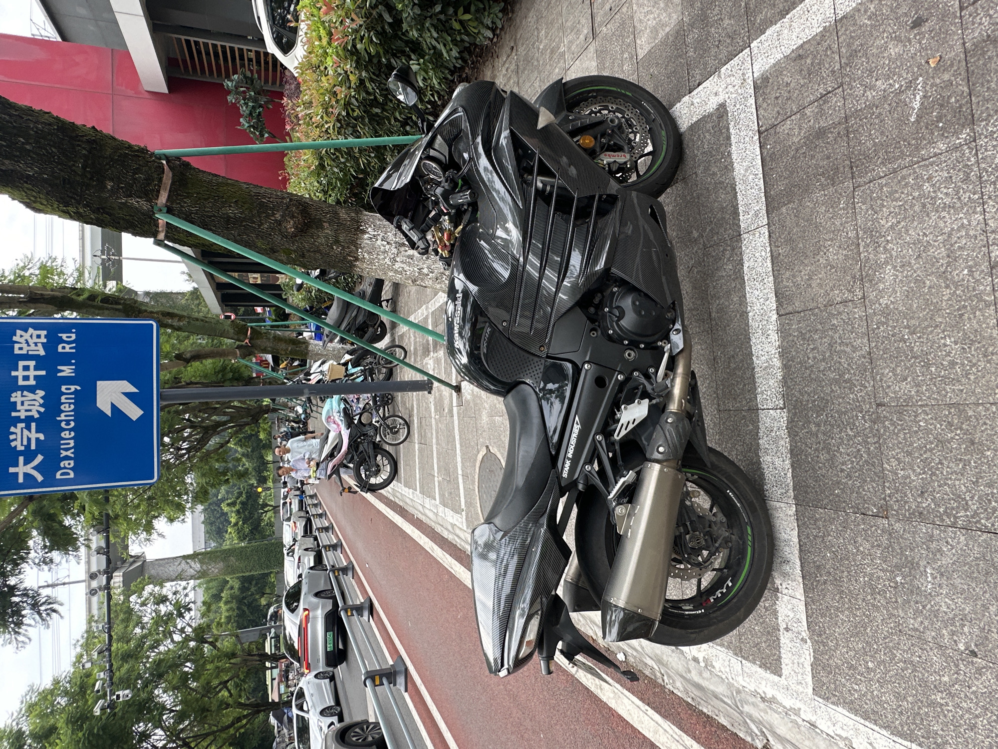A black Kawasaki motorcycle is parked on a sidewalk next to a red bike lane in the Chongqing High-tech Zone, China. The motorcycle is a sportbike with carbon fiber body panels and a large exhaust pipe. It is facing to the right of the frame. A blue sign with Chinese characters and an arrow pointing right is visible above and behind the motorcycle. In the background, several cars and other motorcycles are parked along the street, and a large tree with green supports stands next to the motorcycle. The sidewalk is paved with gray tiles.
