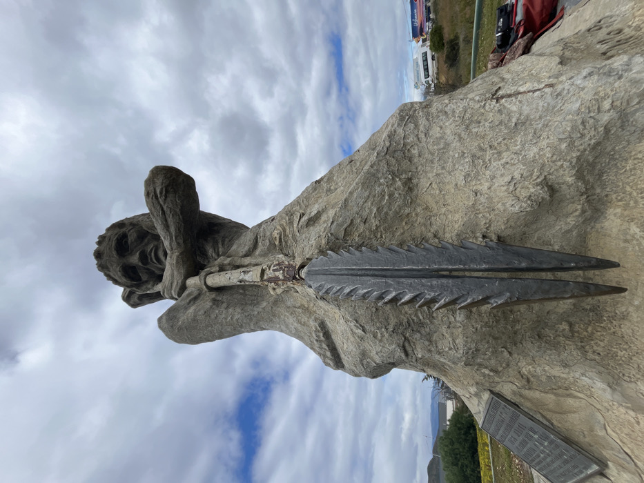 A stone sculpture depicts a reclining figure with furrowed brow and clenched fists resting on a pole topped by a large, double-bladed spear. The figure and spear are integrated into a large, rough-hewn stone base. A plaque sits at the base of the sculpture. The background features a cloudy sky, a road with vehicles, and a hillside with some vegetation. The image is slightly tilted.