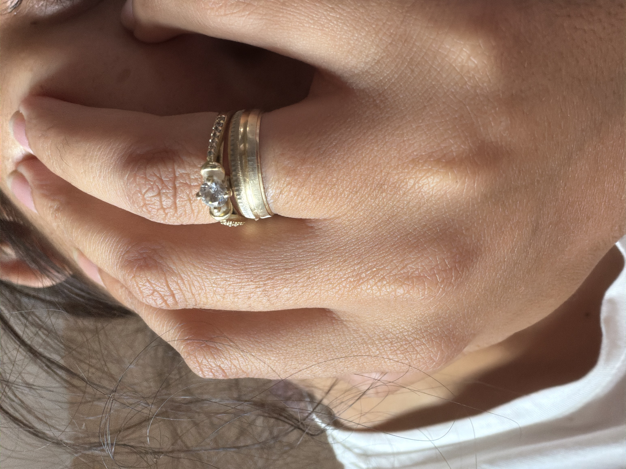 A close-up, medium shot captures a hand adorned with two gold rings, one featuring a small clear stone, gently pressing against a person's face, obscuring part of their eye and forehead. The hand, with visible skin texture and short fingernails, is positioned to the left of the frame, while the person's face, with dark eyebrows and lashes, is visible on the right. Dark hair strands hang loosely around the face and hand. The individual is wearing a white garment, likely a shirt, which is partially visible at the bottom left of the image. The lighting suggests an indoor or shaded outdoor setting. The location in Razhevo Konare, Bulgaria, is noted, but no visual elements within the image directly confirm this geographical context.