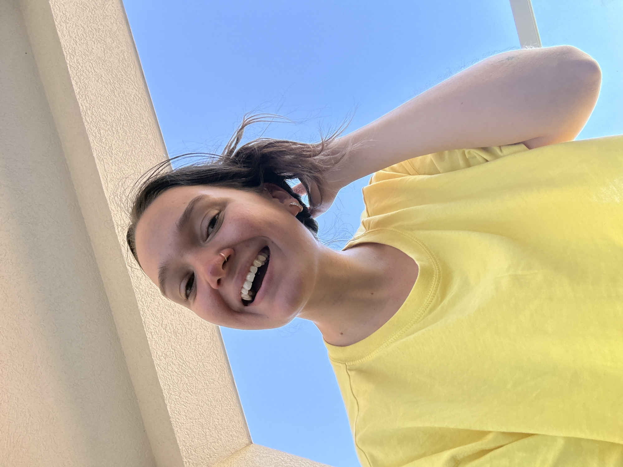 The image is a low-angle, outdoor selfie of a young person wearing a bright yellow t-shirt. The person has dark hair, a nose ring, and is smiling with their mouth open, revealing their teeth. They are touching their hair with their right hand. The background is a clear, bright blue sky. Above the sky is a textured, beige overhang or ceiling. There is a white railing visible on the right side of the image. The scene suggests a sunny day. The location is stated to be Boreti, Montenegro.