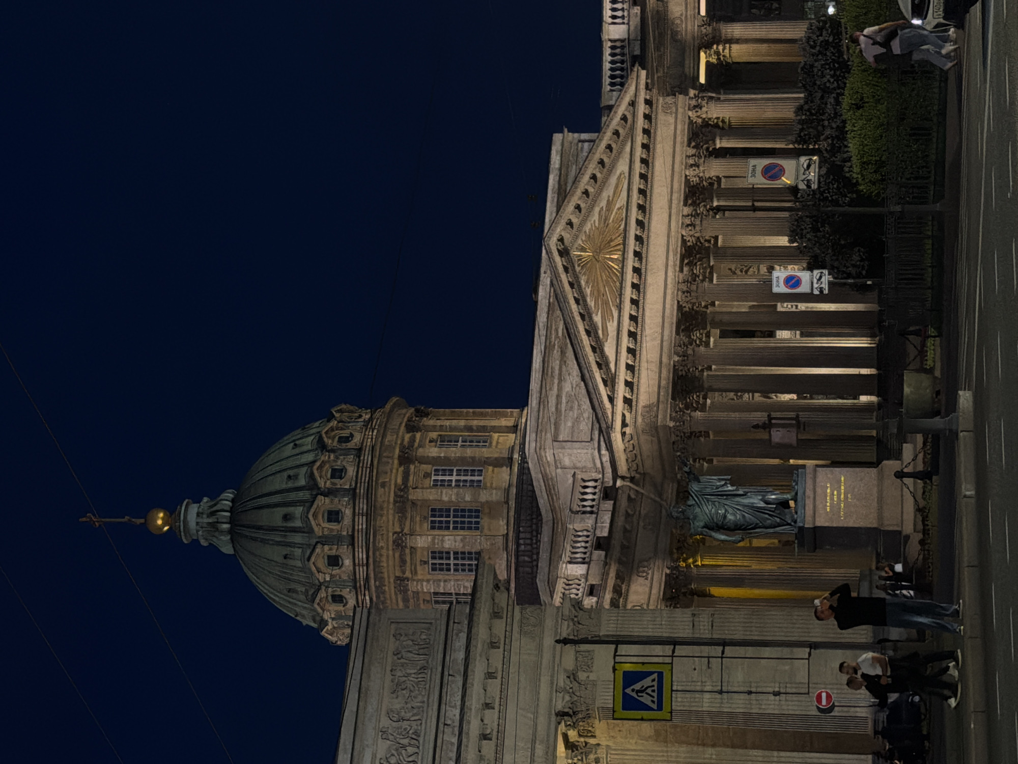 This is a nighttime, medium-shot, eye-level, slightly low-angle photograph of the Kazan Cathedral in Saint Petersburg, Russia. The sky is dark blue and clear, with two thin wires stretching across the upper portion of the frame. The cathedral, made of stone, is illuminated by warm artificial lights, highlighting its classical architecture. The most prominent feature is the large, green dome with a cross on top, surrounded by smaller domed structures. The façade features a colonnade of tall, classical columns and a pediment with a sunburst motif. A bronze statue of a robed figure stands on a pedestal in front of the colonnade.
In the foreground, a paved street is visible with white lane markings. A man in a black long-sleeved shirt and blue jeans stands in the street, holding a phone up as if taking a picture. To his left, two men, one in a white t-shirt and the other in a black t-shirt and dark pants, walk by. A blue pedestrian crossing sign is mounted on a pole to the left of the statue. Further to the right, a group of people are visible in the background near the cathedral entrance. Two women, one wearing a white top and jeans, and the other a dark top and jeans, walk away from the camera towards the street. Behind them, a man in a light pink shirt and jeans, carrying a backpack, walks on the sidewalk. Several cars are parked along the street. The scene is tranquil, with a few individuals captured in moments of pedestrian activity.
