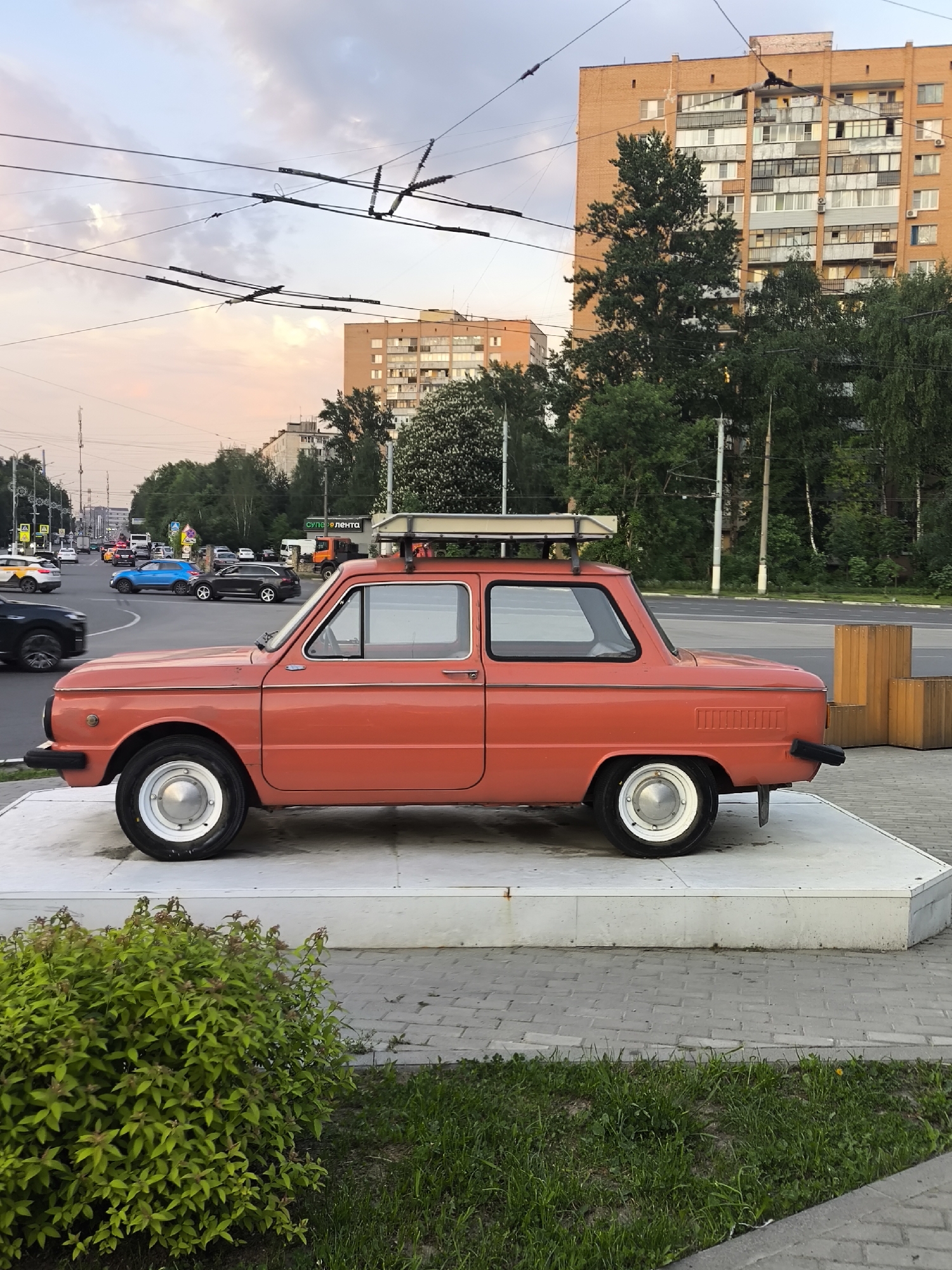 Here's a detailed description of the image:
**Overall Scene:** The image depicts an orange-red car displayed on a concrete platform, likely as an art installation or monument in a city. The background shows a typical urban environment with buildings, trees, roads, and traffic.
**Subject:**
* **Car:** It's an older model, possibly a Soviet-era ZAZ-966 or similar. The car is elevated on a concrete platform. It has a roof rack installed.
**Environment:**
* **Urban Location:** The scene takes place in a city with medium to high-rise apartment buildings visible in the background.
* **Roads and Traffic:** A street with multiple lanes is seen behind the car, including various vehicles.
* **Vegetation:** Trees and bushes are present around the car and along the roadside.
* **Sky:** The sky is partly cloudy with light colors suggesting either early morning or late afternoon.
* **Street infrastructure:** There are traffic lights, overhead electric lines, and what appear to be wooden planters alongside the roadway.
**Objects:**
* **Platform:** A white concrete platform serves as the base for the car.
* **Other Vehicles:** A variety of cars can be seen on the road.
**Location:**
The location is in a Russian-speaking region, specifically a city within the Russian Federation. Signs in the background confirm Russian-language signage. The overall appearance is of a typical provincial city in Russia. The building in the background looks to be a standard Soviet era apartment building.