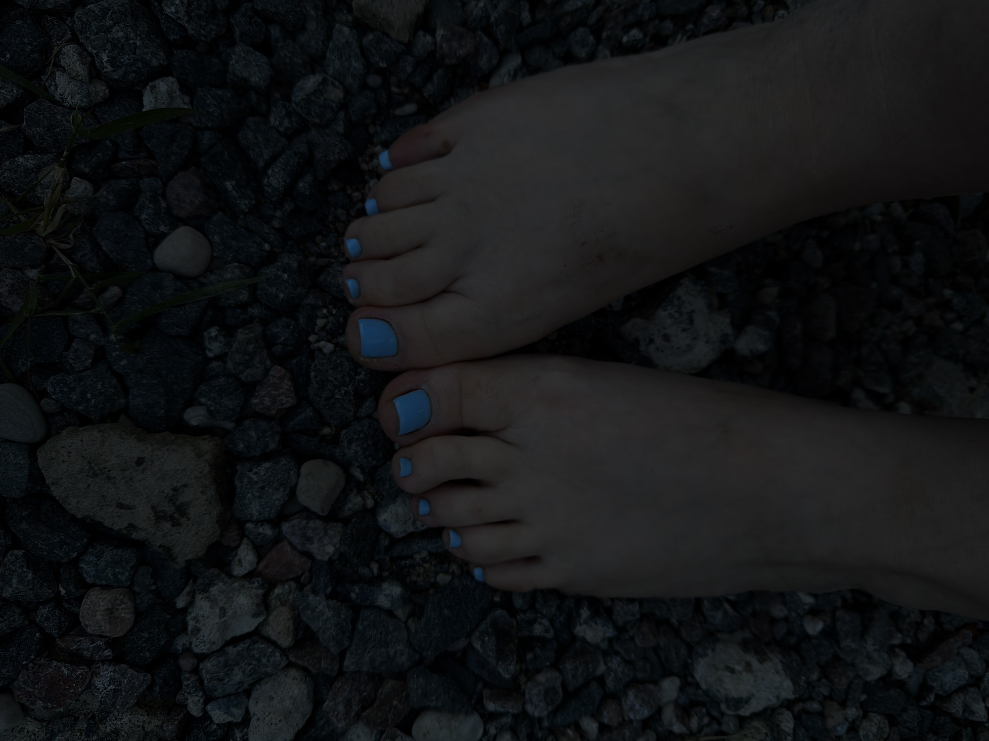 A close-up, overhead shot shows two bare feet with light blue nail polish on all ten toes, resting on a dark, gravely surface. The toes are well-defined, and the skin appears smooth. The background consists of various-sized dark stones and some blades of grass peeking through. The lighting is dim, creating shadows that obscure some of the details of the terrain. No person or action is visible beyond the static placement of the feet.
