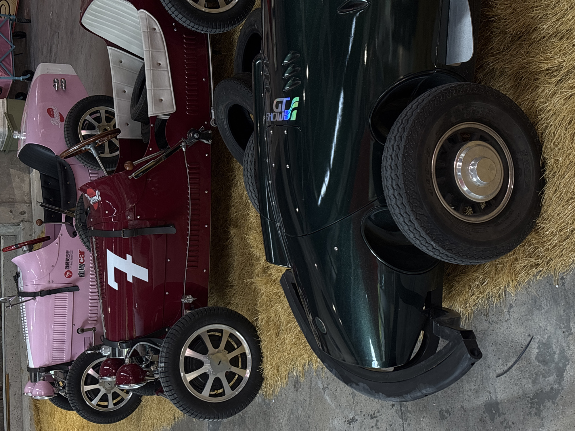 The image shows three vintage-style pedal cars arranged on a bed of dried grass. The car in the foreground is dark green with a chrome hubcap and a black bumper. The middle car is a deep red with a prominent white "7" on its side. The car in the background is light pink and has a white stripe along its hood. The overall setting appears to be an indoor exhibition or showroom. The image does not provide information about the specific location within Pudong, China, or any people or actions.