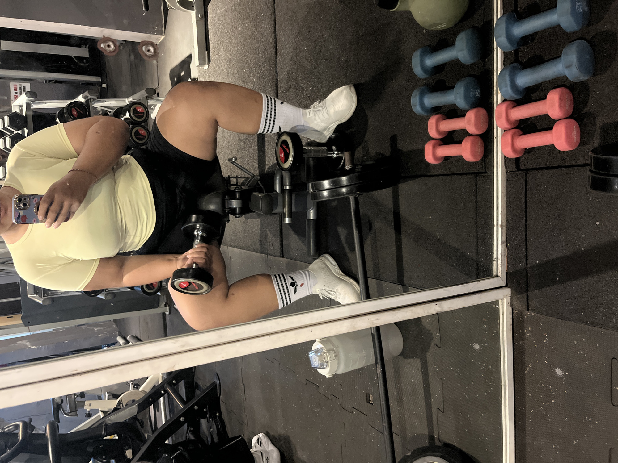 The image depicts a person sitting on a weight bench in a gym, holding a dumbbell in their right hand. The person is wearing a yellow t-shirt, black shorts, white athletic socks with black stripes, and white sneakers. They are also holding a smartphone in their left hand, suggesting they are taking a mirror selfie.
In the foreground, several pairs of dumbbells are visible on the floor. Two pairs are pink, and two pairs are blue. A large, clear water bottle with measurement markings is placed next to the weight bench. In the background, weight racks with dumbbells and other gym equipment are present. A large mirror reflects the scene, allowing for the selfie. The gym floor is black and appears to be made of rubber mats.
The overall environment suggests an indoor fitness facility. There is no specific visual information in the image that can confirm the location in Denpasar, Indonesia.