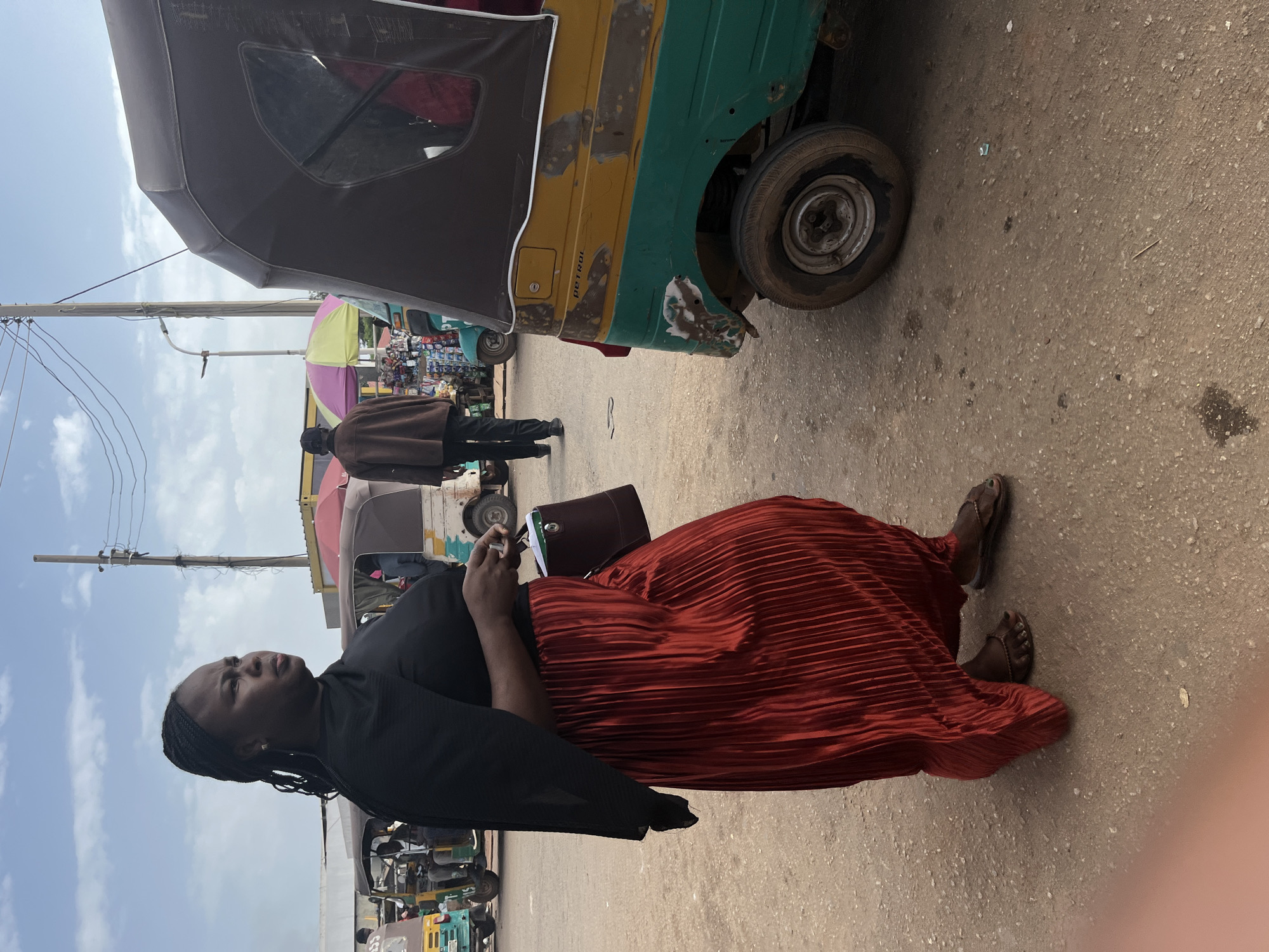 A woman with braided hair stands in what appears to be an open-air market or transportation hub, with several auto-rickshaws and utility poles visible. She is wearing a black top and a long, pleated red skirt. In her right hand, she holds a small dark bag and a white object, possibly a phone or a small notebook. Her gaze is directed to the left. The ground is dusty and uneven. Behind her, auto-rickshaws are parked, and a man in a brown jacket and dark pants walks away from the camera. The sky is partly cloudy. The scene suggests a busy, everyday environment in Gura Top, Nigeria, likely a place where people gather for transportation or commerce.