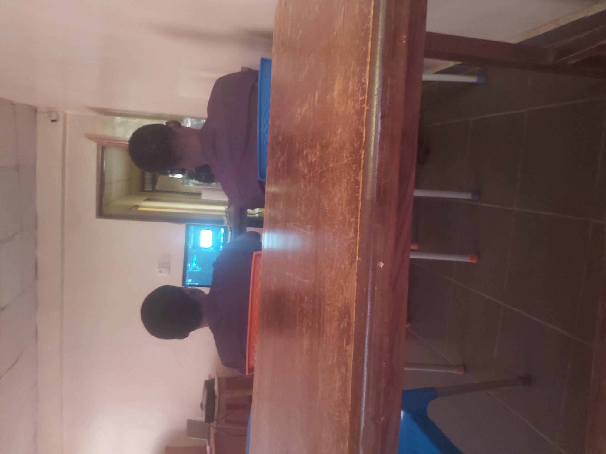 In a classroom or computer lab setting, two individuals, viewed from behind, are seated at a wooden table facing away from the camera. Both are wearing purple shirts. The person on the left is looking at a computer monitor, which displays a bright, blue interface. The person on the right is also facing a computer, but the screen is less visible. The environment appears to be indoors with light-colored walls and a tiled floor. The scene is lit by overhead lighting. The overall impression is one of focused activity, possibly learning or working on computers. The location is identified as Jos, Nigeria.