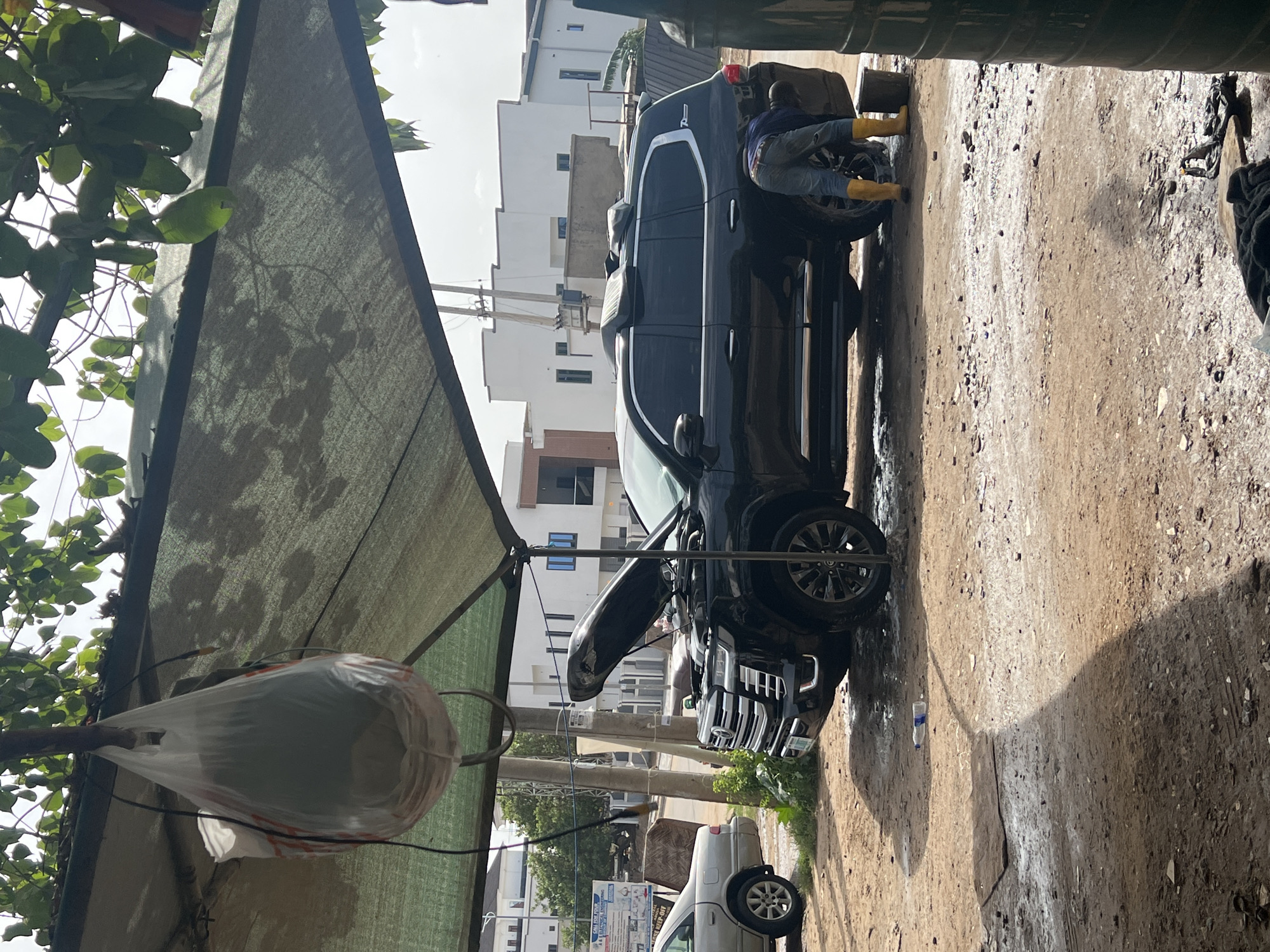 The image shows a vehicle being washed at an outdoor car wash in Guzape, Nigeria. A black SUV is parked on a dirt-covered surface. A person wearing yellow boots is on the side of the SUV washing it. Buildings are visible in the background. A sunshade is present on the left side of the image, with foliage and a plastic bag hanging from it. Another vehicle is parked further down the street.
