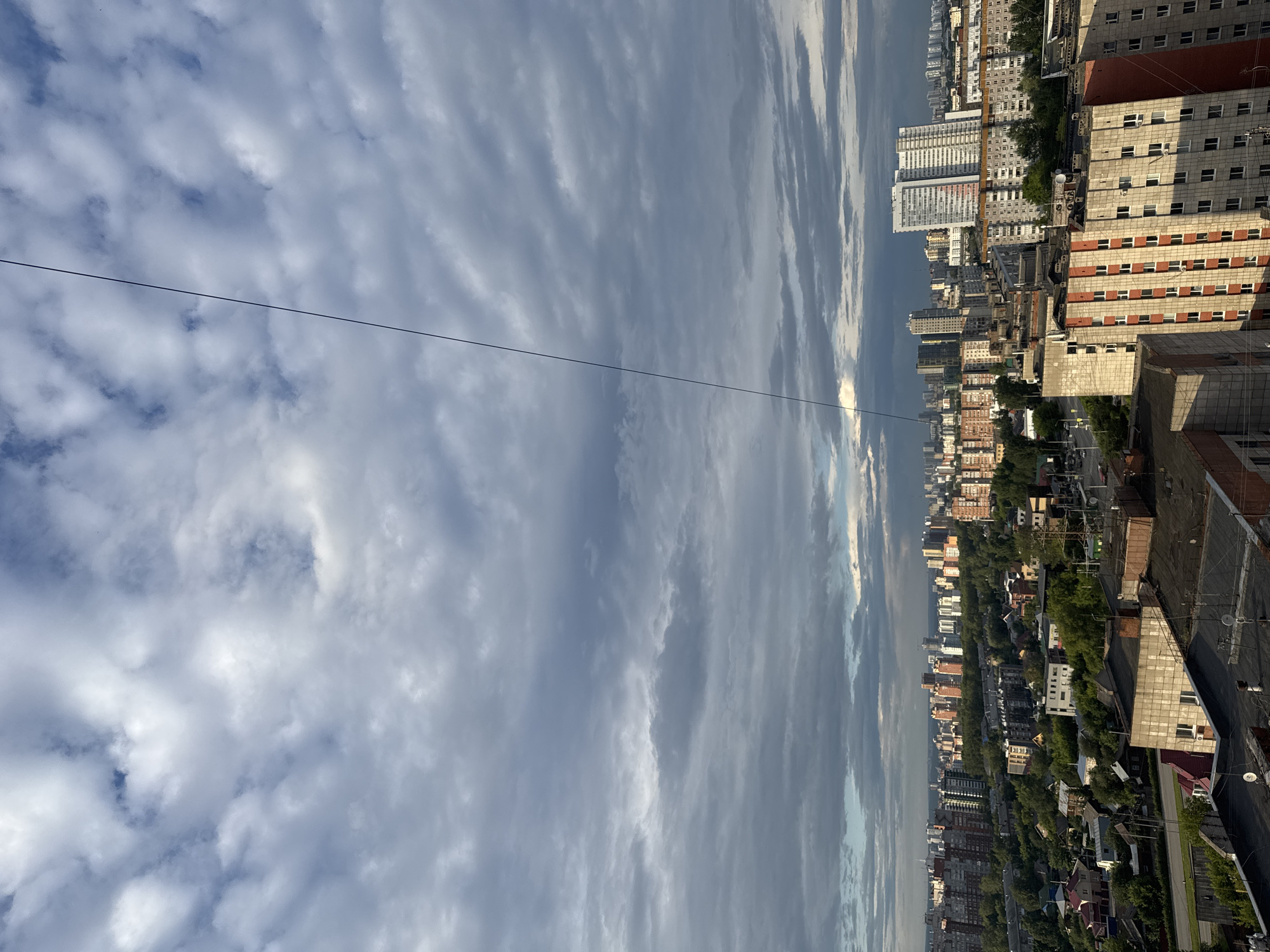 This is an aerial view of a city with numerous residential buildings, a significant amount of greenery, and a cloudy sky. The buildings vary in height and architectural style, with many appearing to be apartment complexes. Some smaller houses with red roofs are visible in the foreground and middle ground, interspersed with trees. A prominent feature is a thin, dark wire extending diagonally from the upper left to the middle of the frame. The city appears to be built on a somewhat hilly terrain. The overall impression is of a densely populated urban area with pockets of green spaces, under a dramatically cloudy sky.
Based on the architectural style of the buildings, specifically the multi-story apartment blocks with balconies and distinct window patterns, and the presence of numerous birch trees and pine trees, this image likely depicts a residential district in the city of Perm, Russia. The layout of the buildings and the urban planning visible are consistent with typical Soviet-era and post-Soviet urban development seen in many Russian cities, including Perm. Without more specific landmarks, it is difficult to pinpoint the exact location within Perm, but the scene is characteristic of its residential zones.