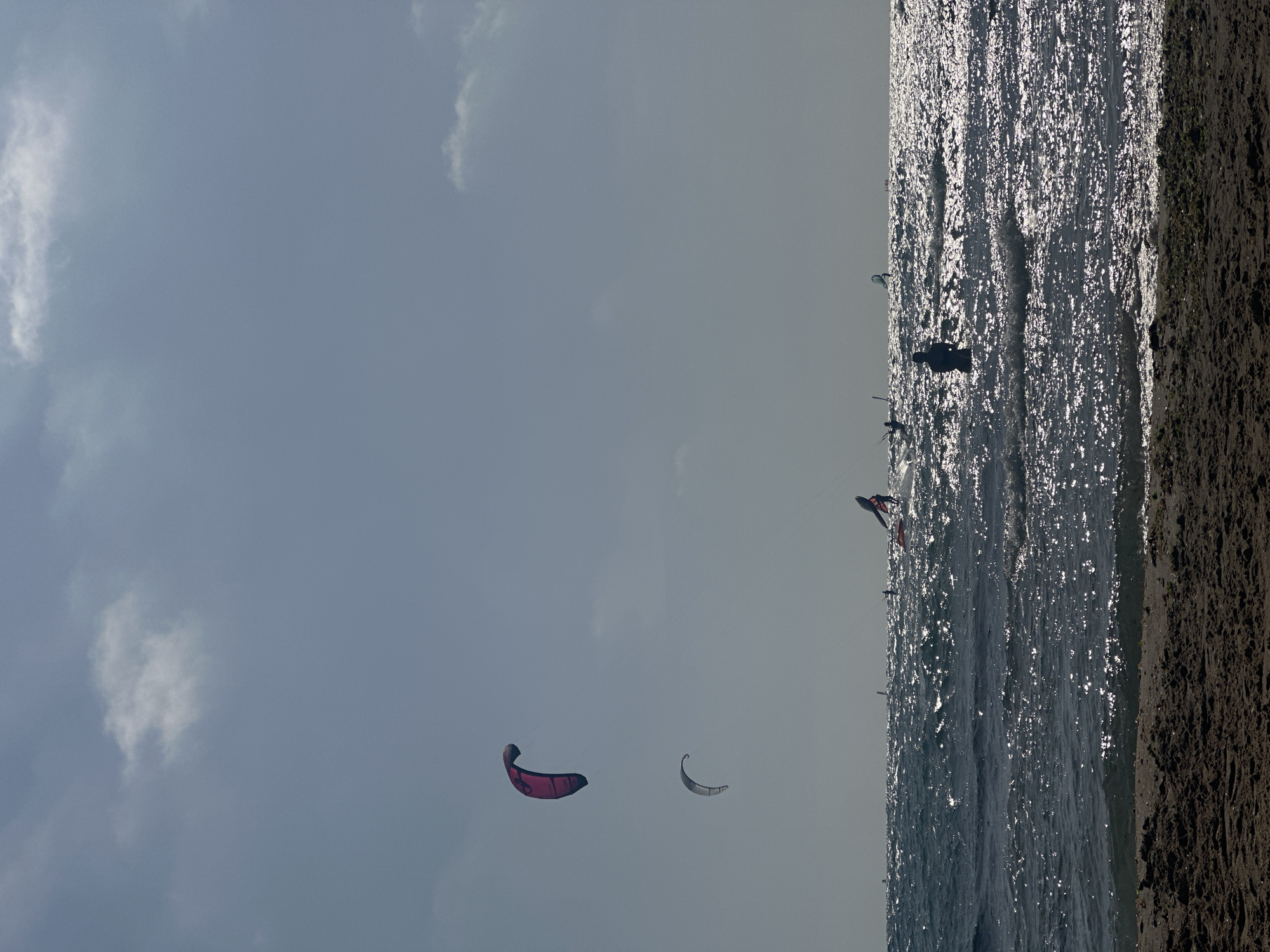 The image captures a beach scene in Tel Aviv, Israel. The frame is oriented vertically, with the sky occupying the majority of the upper portion. Two kites are visible in the sky. The beach runs along the right edge of the frame. The water is choppy, with sunlight reflecting off the surface. Several kitesurfers are in the water, with at least one clearly visible near the center of the frame. The sand is dark and textured.