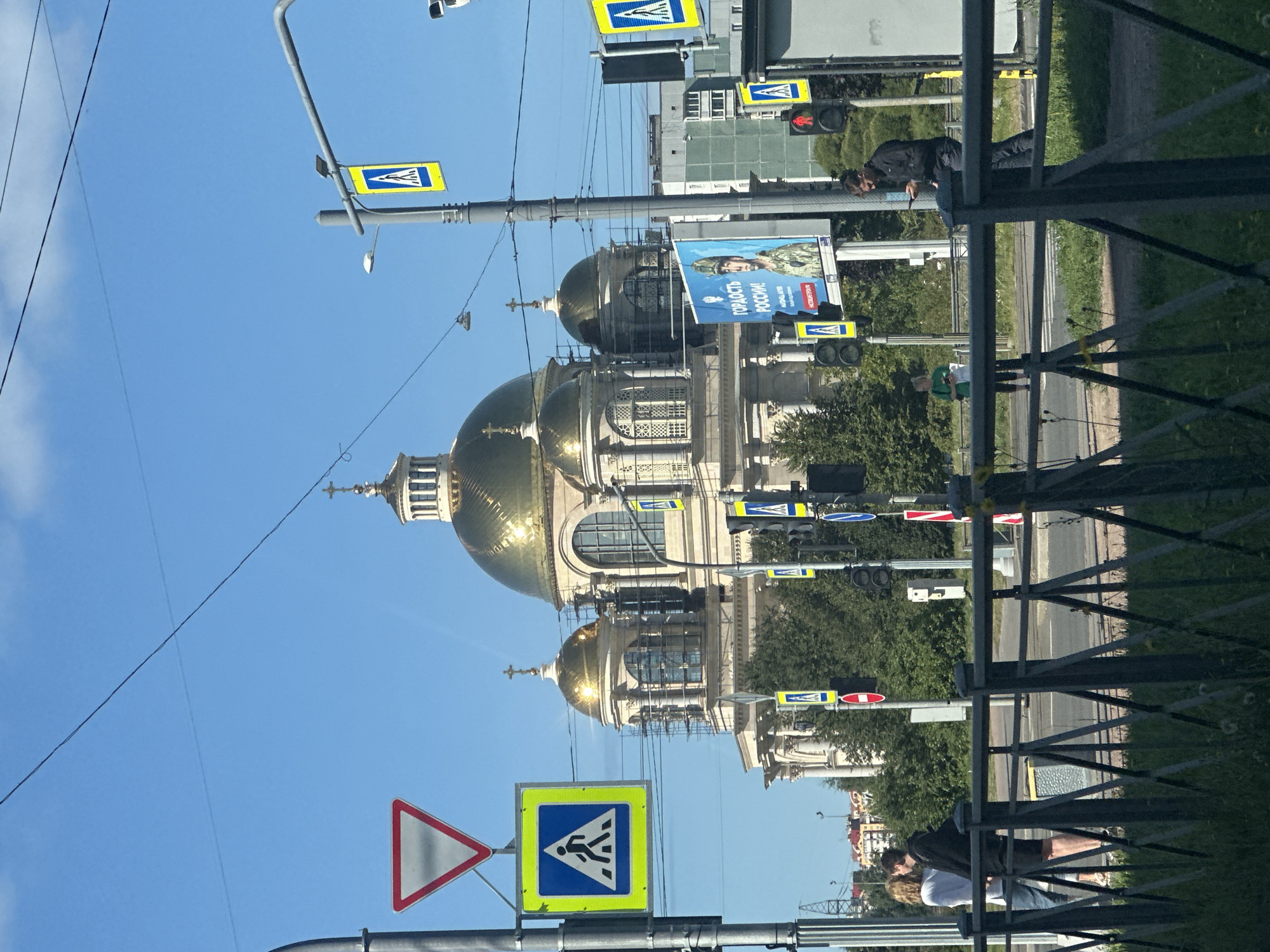 This image shows a street scene in Saint Petersburg, Russia, with a large cathedral featuring a golden dome prominently in the background. The cathedral appears to be undergoing some construction or renovation, with scaffolding visible on its exterior. Several traffic lights and pedestrian crossing signs are present, indicating an urban environment. In the foreground, there is a metal fence with grass and weeds growing through it. People are visible on the sidewalk and crossing the street, including a couple standing close together, a man on his phone, and a woman holding a piece of paper. A billboard displaying a military figure and Russian text is also visible. The sky is clear and blue with some clouds.