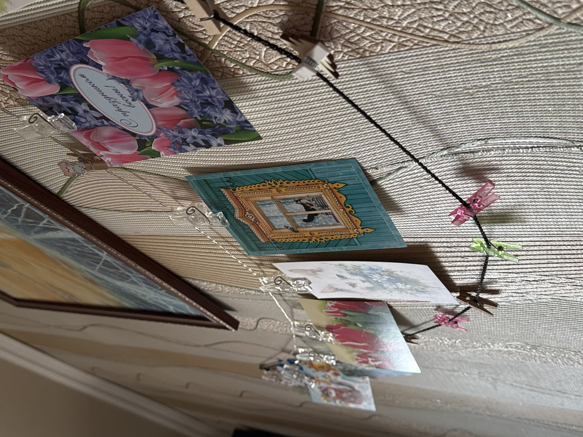 The image depicts a series of postcards and a framed picture hung on a string with small clothespins against a textured, off-white background. The cards display various images, including a floral arrangement with tulips and hyacinths, a scene depicting a window with a cat looking out, and other indistinct landscapes and floral images. The framed picture shows what appears to be a nautical or seascape. The text on the floral postcard is in Russian, reading "С праздником весны!", which translates to "Happy Spring Holiday!". The scene is not explicitly identified with Izhevsk, Russia, but the use of the Russian language on the postcard suggests a connection to the Russian-speaking world.