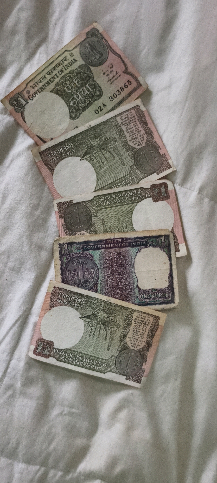 The image shows five Indian one rupee notes and coins, arranged diagonally on a white textured surface. The notes are of various designs and appear to be from different years, some featuring the Ashoka pillar and text in Hindi and English. Several notes have circular white watermark-like impressions. The coins appear to be one rupee coins, with one coin visible on each of the top four notes. The lighting is dim, suggesting an indoor setting, possibly a bedspread. No people are visible in the image. The scene is a close-up shot of currency notes and coins. There is no indication of the city of Kundrathur, India, or any urban environment in the image.