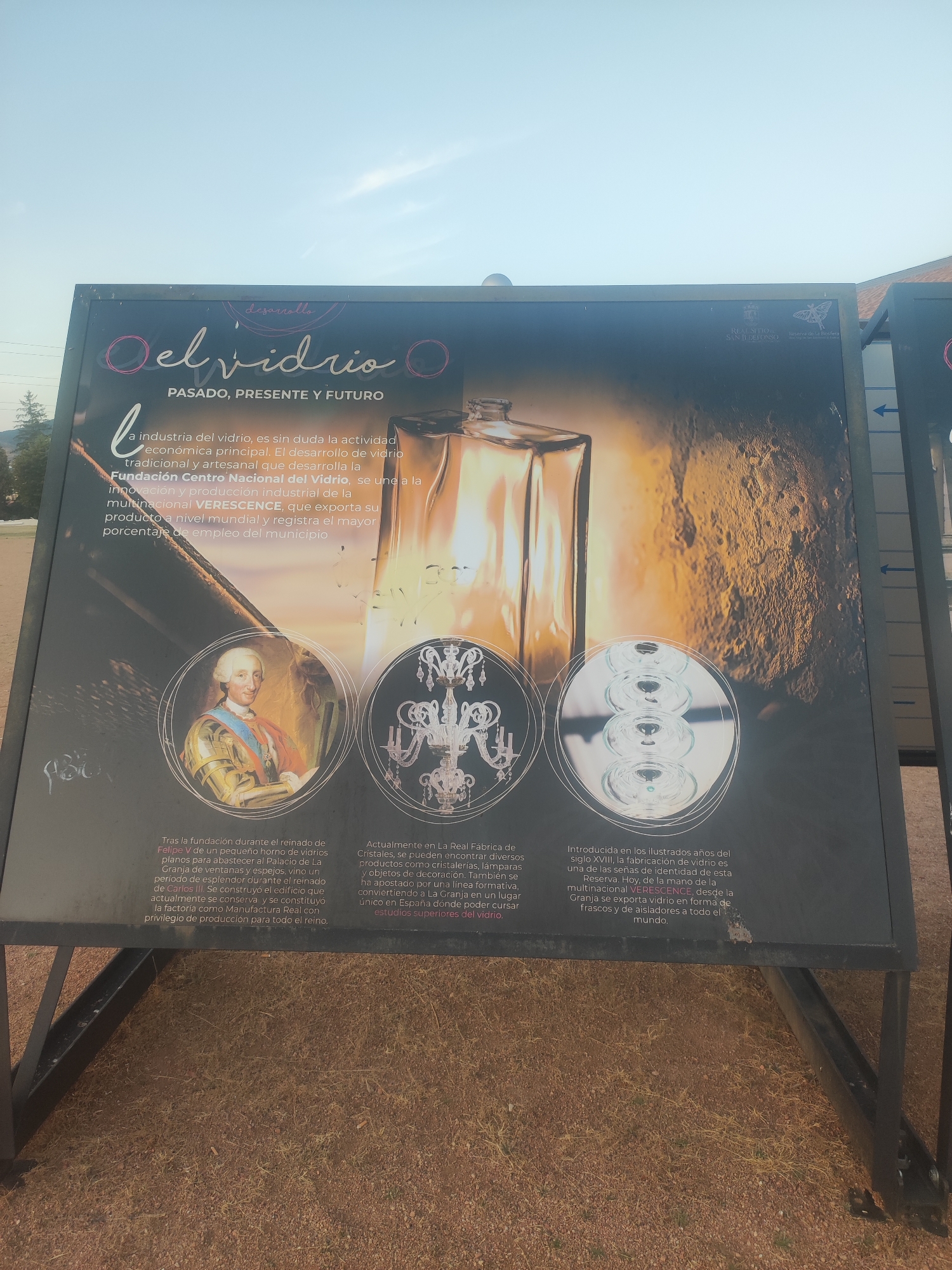 The image shows a large outdoor information panel in the city of Real Sitio de San Ildefonso, Spain. The panel is black with text and images related to the history and present of glassmaking. The main title reads "el vidrio PASADO, PRESENTE Y FUTURO" (glass PAST, PRESENT AND FUTURE). There is an illustration of a glass bottle, a portrait of King Ferdinand VI, a chandelier, and several glass lenses. The text on the panel describes the importance of the glass industry in the region and its historical significance, mentioning the founding of a glass factory during the reign of Ferdinand VI and its continued operation. The panel is set up outdoors, with the ground appearing to be covered in dirt and dry grass. The sky is visible in the upper part of the image.