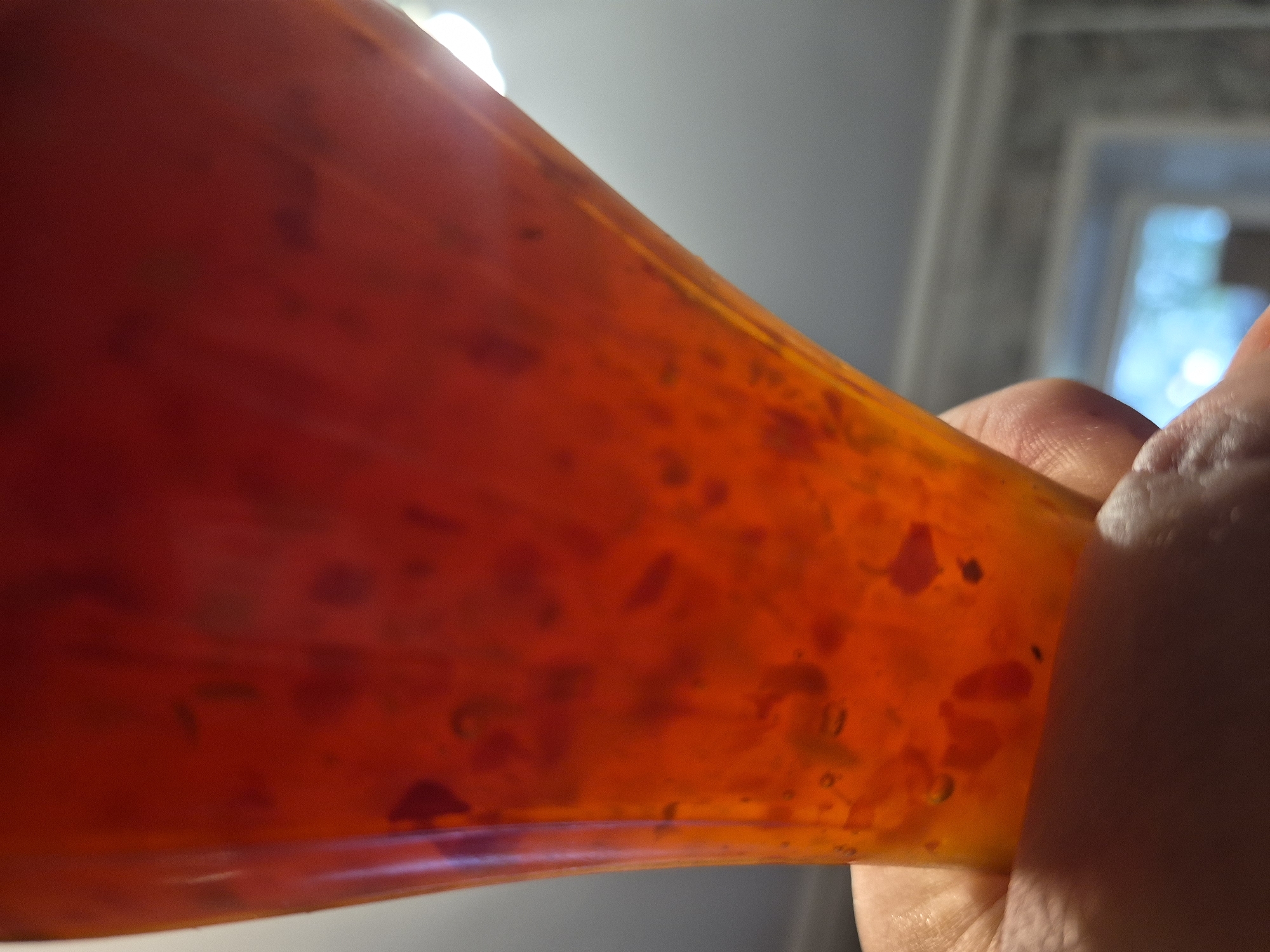 The media file shows a close-up, low-angle shot of a hand holding a translucent, orange-red colored substance. The substance appears to be some sort of gel or viscous liquid, with darker red flecks or inclusions suspended within it. The light source is strong and appears to be coming from behind and above, causing the material to glow and highlight its translucent nature. The hand is partially obscuring the right side of the object, with fingers visible. The background is out of focus and appears to be an indoor setting with diffused natural light coming from a window or opening, possibly indicating daytime. There are no visible signs of text in the media file. The overall impression is abstract, focusing on the texture, color, and light interaction of the substance being held. The setting is not clearly identifiable beyond being indoors, and there are no specific details to pinpoint the location within Nyagan, Russia.