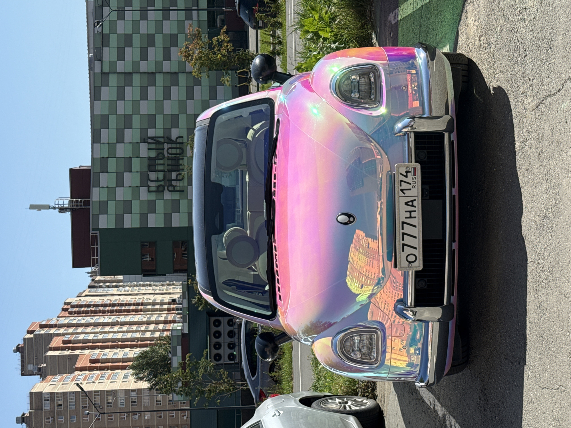 A holographic pink car is parked on asphalt in front of a building with a green checkered facade. The car has a license plate that reads "O 777 HA 174 RUS". Behind the car is a sidewalk with green painted lines and grass. In the background, there are several multi-story buildings, including one with a sign in Russian that says "БЕЛЫЙ РЫНОК" (White Market). The sky is clear and blue. There are no people in the immediate vicinity of the car.