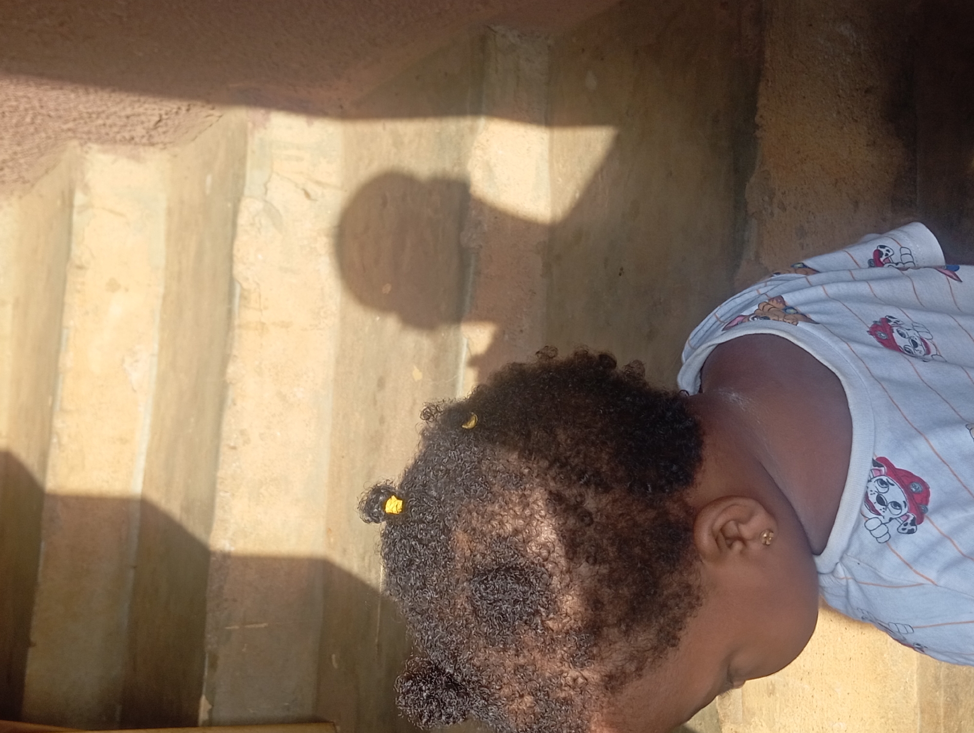 The image provides a close-up, high-angle view of a young child, likely a toddler, in Nkpor, Nigeria. The child is lying on a light-colored, textured ground surface, positioned next to a series of worn, light brown concrete steps or risers.
The subject is a child with dark, curly hair, adorned with two small yellow hair ties or clips. One ear is visible, featuring a small gold stud earring. The child is wearing a white short-sleeved t-shirt with thin, light-colored stripes and various colorful cartoon dog characters, identifiable as characters from Paw Patrol (e.g., Marshall). The child's posture suggests they are resting or possibly sleeping.
The scene is brightly lit by direct sunlight, casting strong, defined shadows across the steps and the ground. A distinct shadow resembling a human head is visible on the wall adjacent to the child, indicating the presence of another person, likely the photographer. The intensity and angle of the shadows suggest it is either morning or late afternoon. The overall setting appears to be an outdoor or semi-outdoor area, consistent with a casual or domestic environment in a sunny climate. No discernible text is present other than the patterns on the child's clothing.