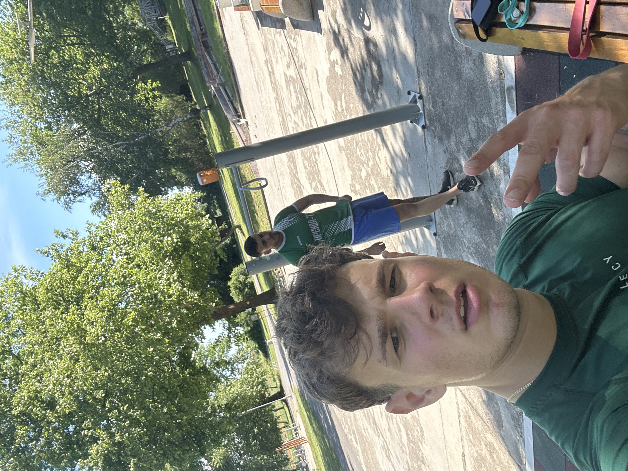 The image is a selfie taken outdoors, possibly in a park setting in Elhovo, Bulgaria, based on the text on the t-shirt of the other individual in the photo. The primary subject is a young man wearing a green t-shirt. Another young man is further away in the background, wearing a green and white t-shirt and blue shorts. He is standing near a metal exercise station. In the background, there are trees and a concrete surface that appears to be a walkway or a park area. Part of a wooden bench is visible in the upper right corner, and some exercise equipment is nearby. The selfie taker has a finger pointing towards the other individual.