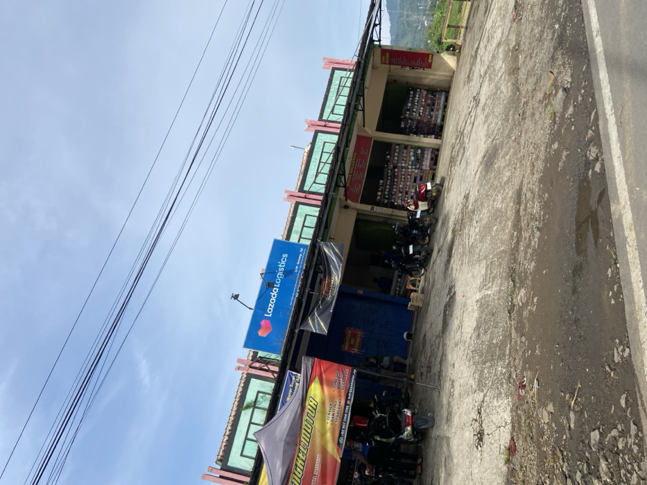 The image shows a storefront with a sign displaying "Lazada Logistics" on the front. The facade of the building is made of concrete with green and pink trim. There are two awnings over the entrance, one of which has a sign that reads "Angel Motor". The building is located in Soreang, Indonesia, at the coordinates -7.035, 107.526.  There are multiple motorcycles parked in front of the store. Several people are standing near the entrance, interacting with the merchandise. The ground in front of the store is made of concrete and has a small puddle of water. There is a small patch of grass in front of the store, and in the background, one can see a hilly, forested area.  A blue sky with some clouds is visible in the upper portion of the frame.  Several electrical power lines crisscross the sky.  The image was taken during the daytime.