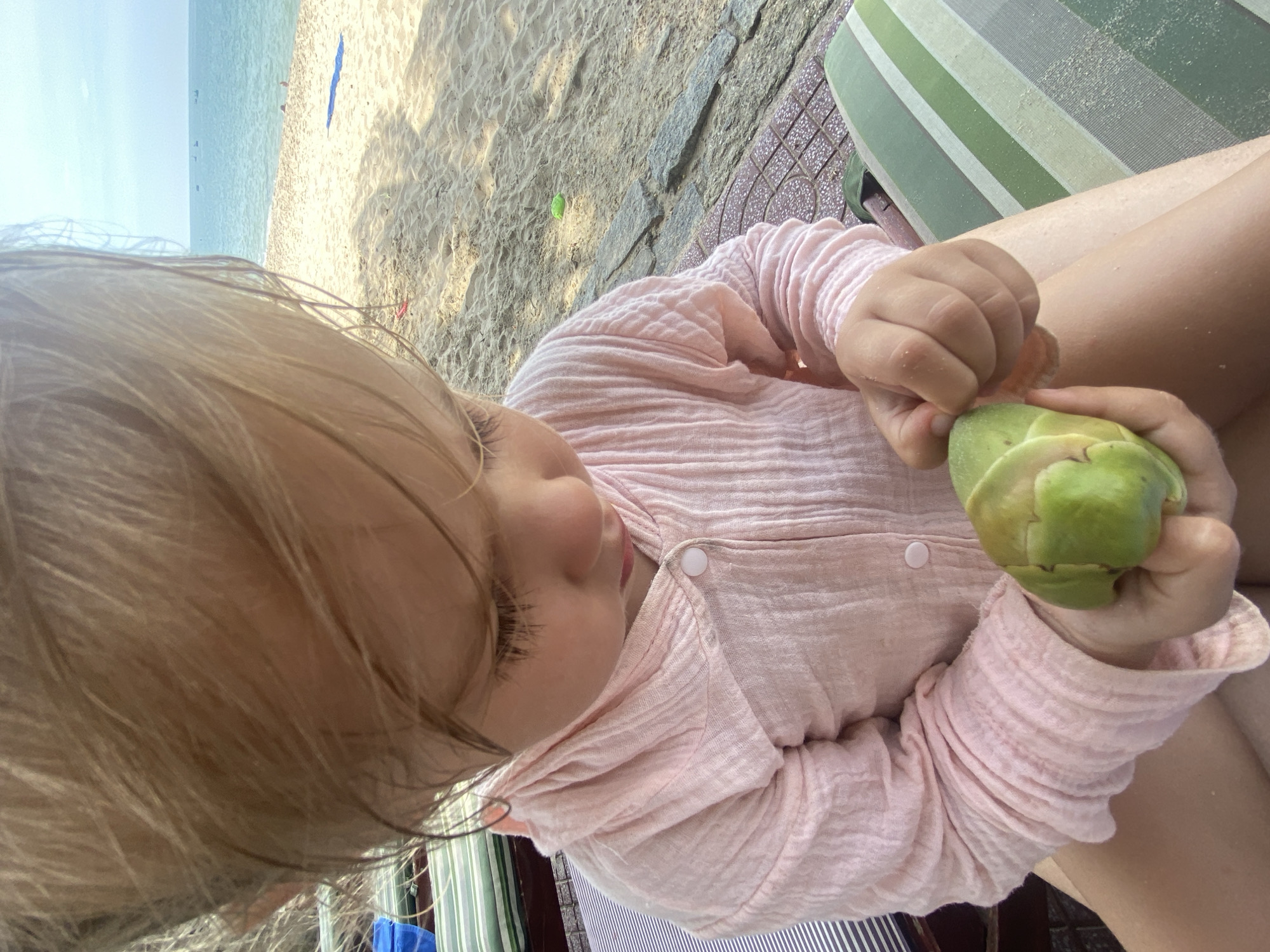 A young child with blonde hair sits on a striped lounge chair on a beach in Phu Thuy Ward, Vietnam. The child is wearing a pink hooded shirt with buttons and is looking down at a green, leafy object held in their hands. The beach has sand and small rocks, and the ocean is visible in the background with a few boats on the horizon under a clear sky.