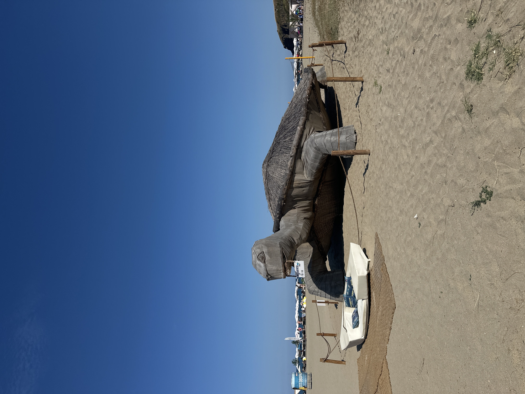 The image shows a large, man-made turtle structure on a sandy beach under a clear blue sky. The turtle is situated in the foreground, with its head facing left. Its shell is thatched with straw-like material, and the body appears to be covered in a grey fabric. In front of the turtle is a white, modular sofa with dark blue cushions, on which the word "RESERVED" is printed in a repeating pattern. Several wooden posts with rope are placed around the turtle and sofa.
In the background, the beach extends towards the sea, with rows of white umbrellas and lounge chairs visible. There are a few people scattered on the beach. To the left, a blue sign with white lettering is partially visible, and a beach volleyball net is present further back. The coastline curves to the right, with a grassy, rocky hill in the distance. The location is identified as a beach in Sinemorets, Bulgaria. The scene appears to be a resort or a leisure area on a sunny day.