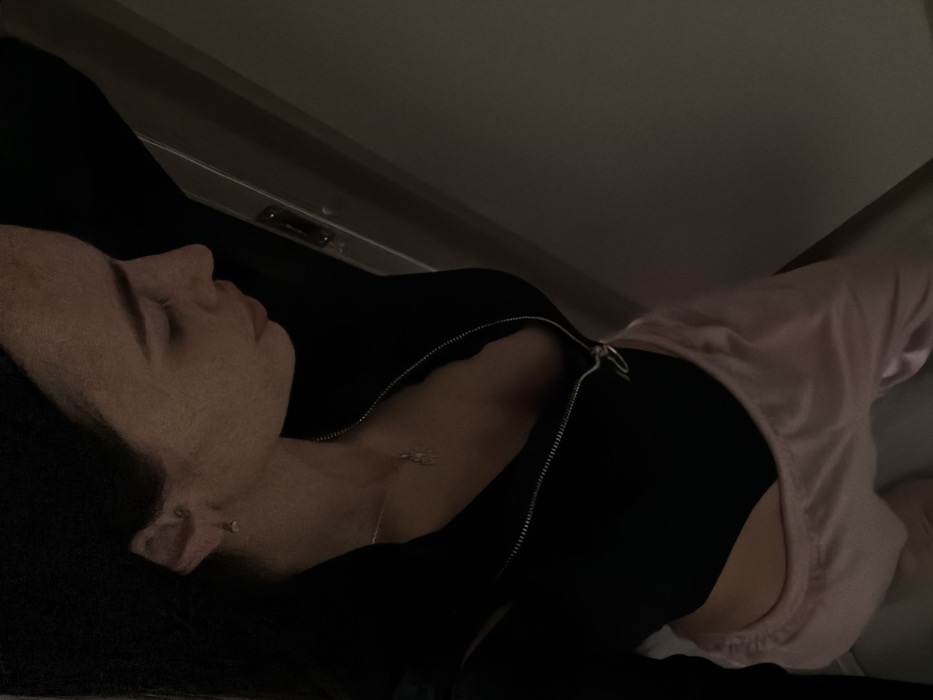 A dimly lit, high-angle interior photograph displays a female individual, located in Krasnaya Zarya, Russia. The subject is depicted from the mid-torso upwards, positioned in profile, with her dark hair falling behind her head. Her gaze is directed downwards and towards the right side of the frame. She is wearing a long-sleeved black top with a silver zipper, partially unfastened to expose the upper chest. Beneath the top, the waistband of light pink, loose-fitting pants, possibly satin, is visible. A delicate silver-colored necklace with a small, indistinct pendant is around her neck, and a small earring is present in her visible ear. Her right arm is raised, with the hand out of frame, suggesting she is leaning against an unseen vertical surface. The background consists of plain, light-colored walls, with a portion of a doorframe or architectural panel, possibly including a metallic latch mechanism, visible to the right of her head.