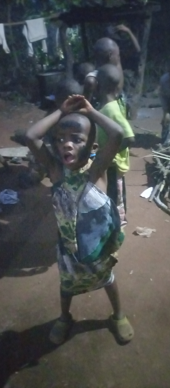 This image depicts a low-light, likely evening or nighttime, scene in Amokwue-Affa, Nigeria, featuring several children in a rustic outdoor setting.
The main subject is a young child in the foreground, facing the camera with a dramatic, wide-eyed expression and open mouth, hands raised above their head. This child wears a patterned dress or tunic, a mix of green, black, and white, and light-colored slip-on shoes. Dark markings, possibly paint or dirt, are visible on their face.
Behind the central figure, other children are present, including one in a light green shirt looking away from the camera. The ground is bare earth or dirt. In the dimly lit background to the left, white fabric, possibly laundry, hangs on a line or branch, near a large dark pot or container and some bottles. To the right, a pile of sticks or cut wood is visible. The overall scene suggests a candid moment of play or activity in a village environment. No text is visible in the image.