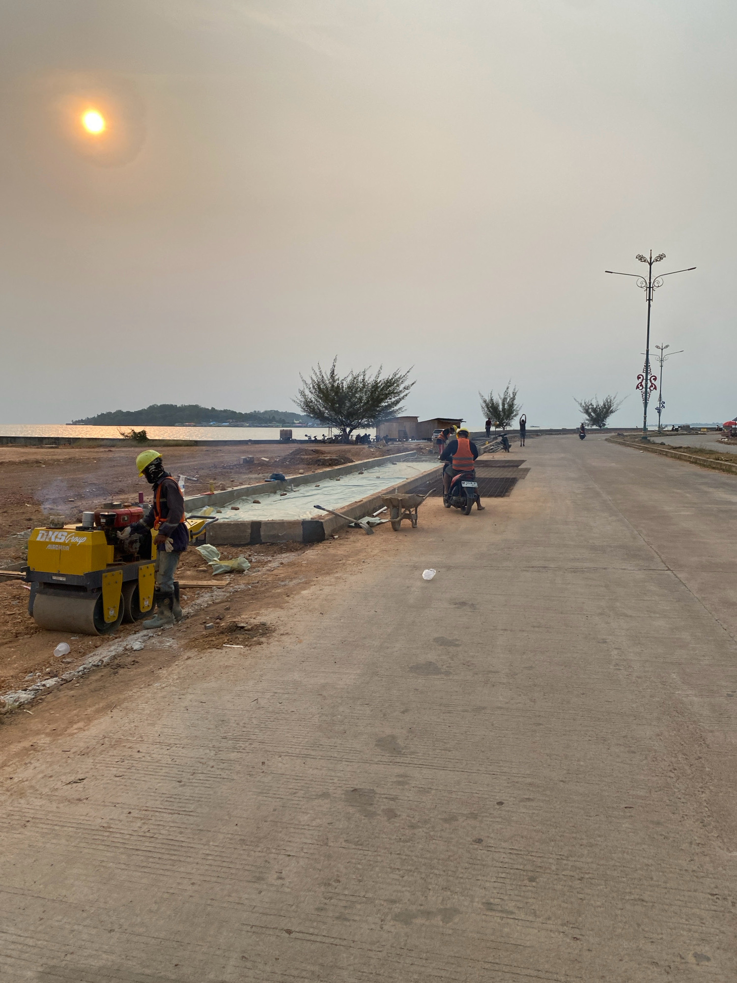 The image captures a scene of road construction in Tanjung Pinang, Indonesia, likely near the waterfront given the presence of the sea and a distant island. The sky is hazy with the sun appearing as a bright, diffused orb in the upper left quadrant. In the foreground, a construction worker in a yellow hard hat and an orange safety vest operates a yellow vibratory roller on a dirt and gravel surface. The roller is compacting a new section of roadbed that has been covered with a light-colored material, possibly geotextile fabric, and has concrete curbs being formed.
Further into the scene, other construction workers are present, with some operating a motorcycle. One individual on the motorcycle wears an orange safety vest. A wheelbarrow is visible to the left of the new road section. In the background, the sea stretches out, reflecting the hazy sunlight. Several sparse trees with twisted trunks dot the landscape between the construction site and the water's edge. Along the right side of the image, a newly paved concrete road extends into the distance, flanked by decorative streetlights. Several motorcycles and people are visible further down this road, suggesting ongoing activity or a public area. The overall impression is one of ongoing infrastructure development under hazy atmospheric conditions.