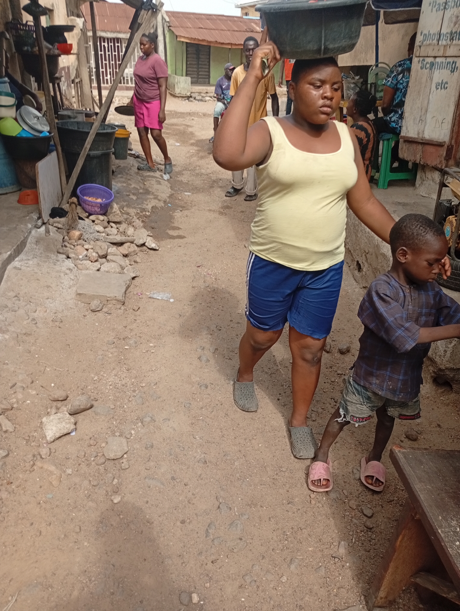 In Adankolo, Nigeria, a dirt lane reveals a scene of everyday life. A woman in a yellow tank top and blue shorts walks toward the camera, carrying a dark basin on her head. A young boy in a plaid shirt and tattered shorts walks beside her. Behind them, other people are visible in the distance. Various buildings and structures flank the lane, including what appears to be a business providing passport photos and scanning services. The ground is uneven and rocky.