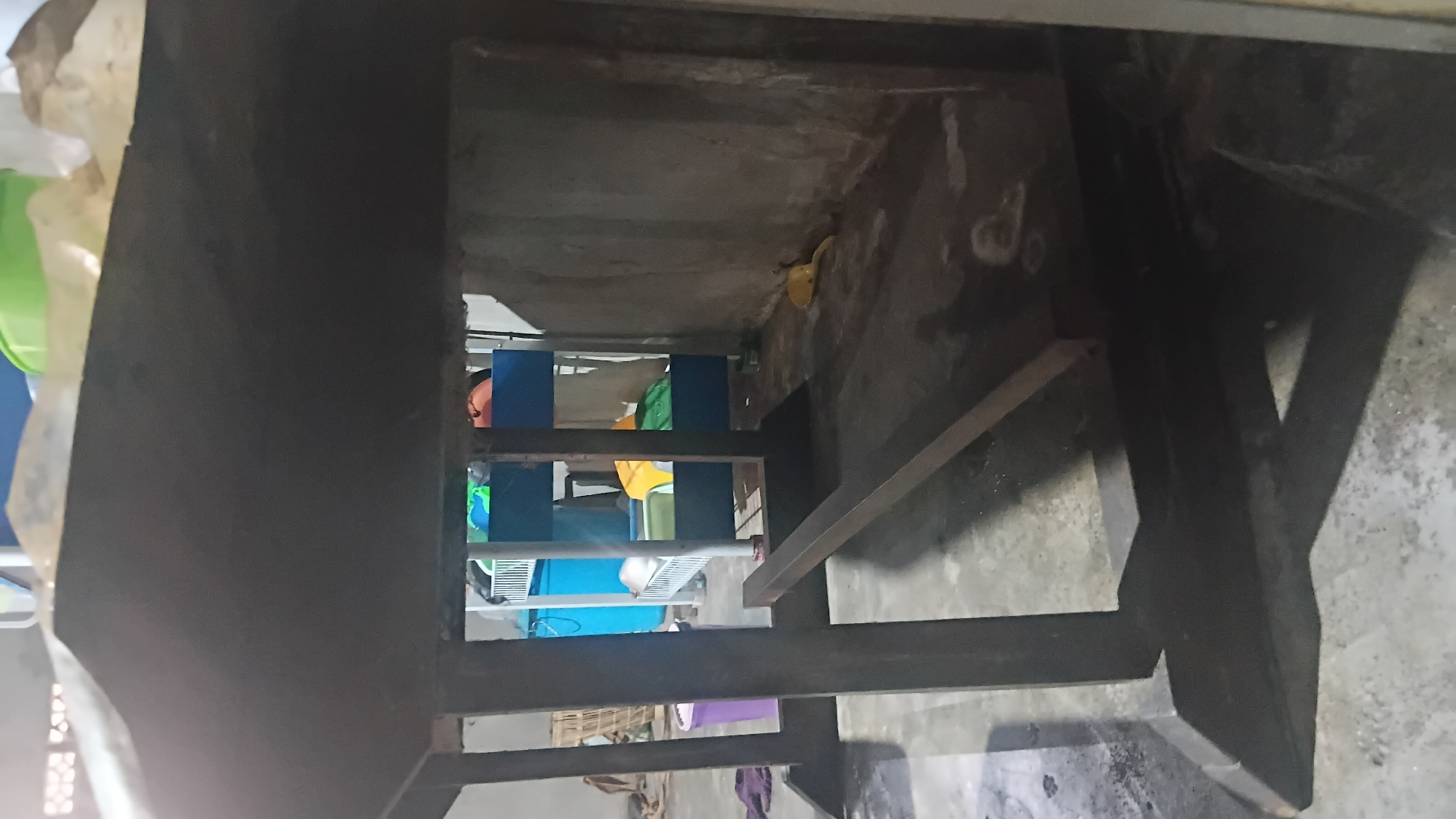 A metal table frame is in the foreground of this image, with an opening revealing a cluttered indoor space in the background. The scene appears to be a storage area or workshop, possibly located in Prabumulih, Indonesia. The table frame is made of dark, thick metal beams and has a raw, unfinished appearance. The opening in the frame offers a view into a room with a concrete floor and walls. Behind the table, a multi-tiered shelving unit is visible, holding various items including blue and green plastic containers, a yellow object, and possibly clothing. To the left of the shelving unit, a woven basket and a purple trash can are present. The lighting is dim, casting shadows and obscuring some details. The overall impression is one of a functional, yet unorganized, space.