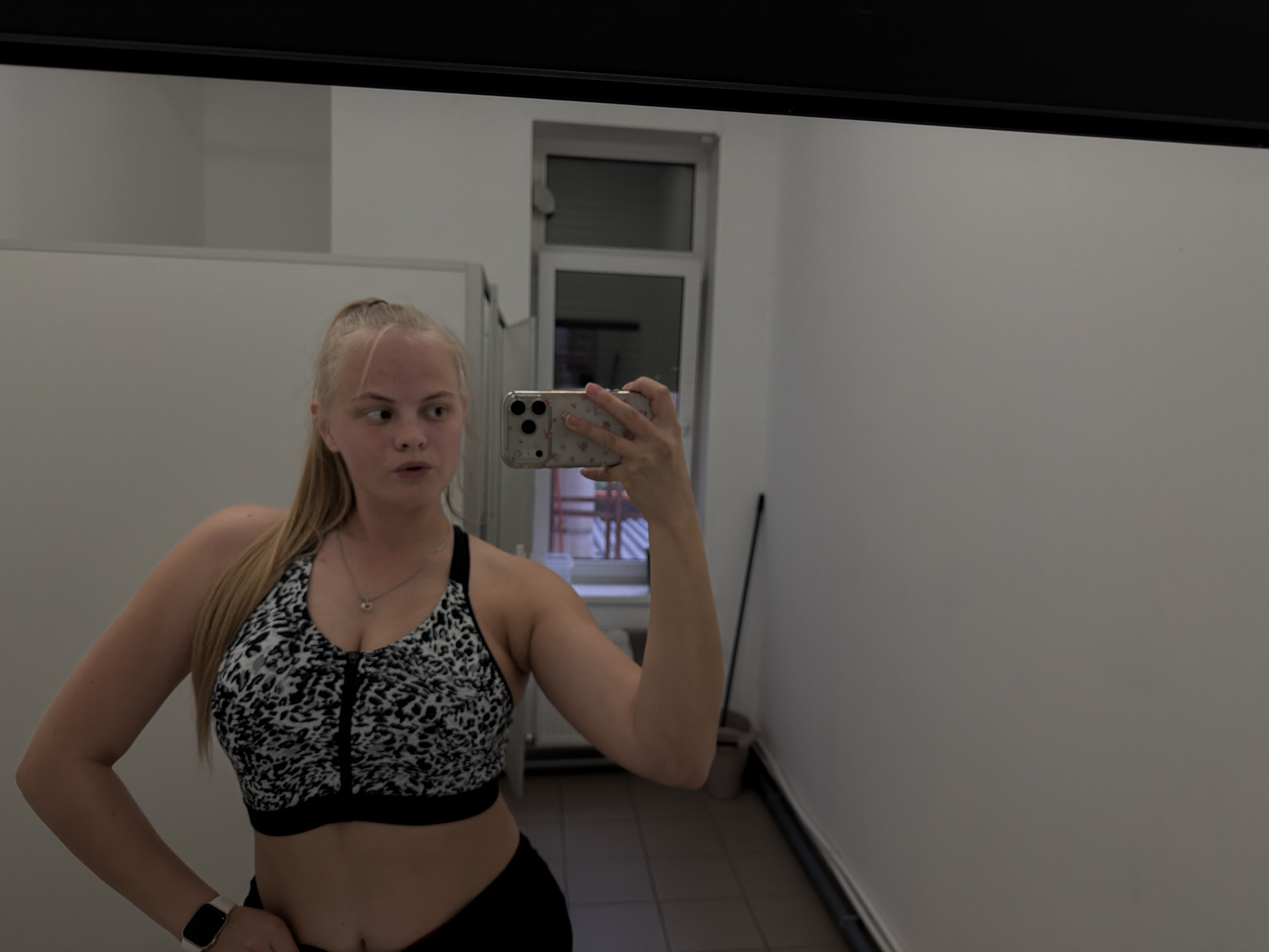 A young woman with blonde hair pulled back in a ponytail is taking a selfie in a bathroom mirror. She is wearing a black and white sports bra and black athletic pants, and an Apple Watch on her left wrist. The bathroom has white walls and a dark-framed window. There's a tall white cabinet to her left and a trash can near the window. No specific location in Izhevsk, Russia can be identified from the image.