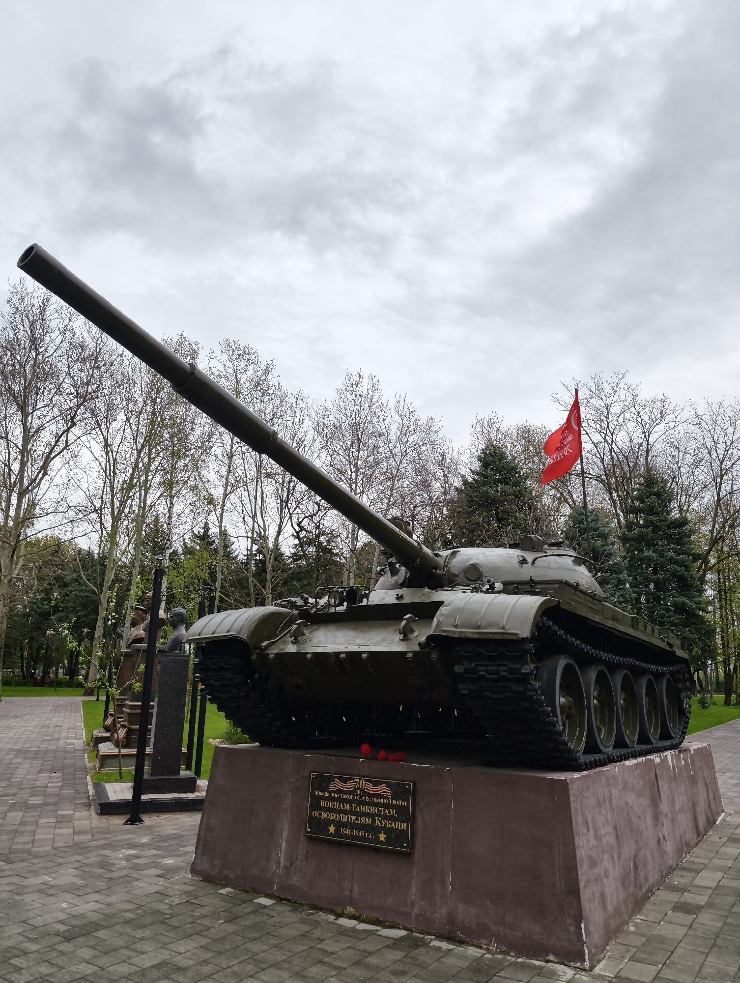 A large, olive-green tank, likely a Soviet-era model such as a T-54/55, serves as the central subject of this memorial in Krasnodar, Russia. The tank is positioned on a dark brown, tapering pedestal, angled slightly upwards with its long barrel pointing towards the left of the frame. The tank features robust tracks, visible road wheels, and various hatches and equipment on its turret and hull.
To the right of the tank, a red flag with white Cyrillic text, partially legible as "ЗА..." (likely "За Родину!" - "For the Motherland!" or a similar patriotic slogan), flutters from a pole, adding a splash of color to the otherwise muted palette. Behind the tank and flag are dense evergreen trees, while sparser deciduous trees with bare branches stand further back and to the left, suggesting early spring or late autumn.
In the mid-ground, to the left of the tank, several dark busts on pedestals are visible along a paved walkway. The ground around the memorial is covered with well-maintained green grass and a patterned paved path. The sky is overcast and grey, indicating a cloudy daytime setting.
A plaque affixed to the pedestal beneath the tank reads in Cyrillic:
"70 ЛЕТ ПОБЕДЫ В ВЕЛИКОЙ ОТЕЧЕСТВЕННОЙ ВОЙНЕ" (70 YEARS OF VICTORY IN THE GREAT PATRIOTIC WAR)
"ВОИНАМ-ТАНКИСТАМ," (TO TANKER-SOLDIERS,)
"ОСВОБОДИТЕЛЯМ КУБАНИ" (LIBERATORS OF KUBAN)
"* 1941-1945 г.г." (1941-1945, years)
Small red flowers are placed on the pedestal above the plaque, likely commemorating the event. The scene conveys a solemn atmosphere befitting a war memorial.
