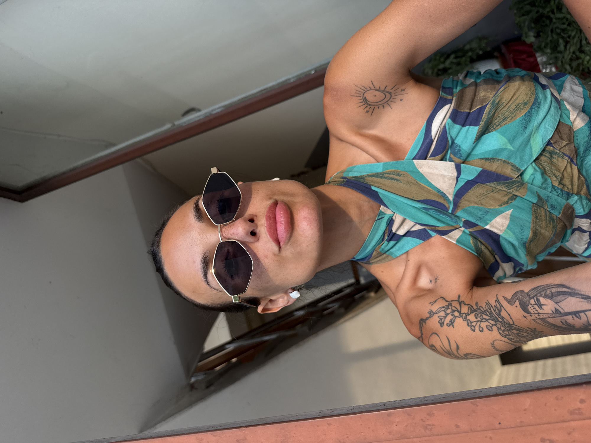 A young woman is pictured from the chest up, looking towards the viewer. She is wearing gold-rimmed, angular sunglasses and a halter top with a blue, green, and brown abstract leaf pattern. Her hair is pulled back, and she has small white earrings. Tattoos are visible on her left arm and shoulder, including a sun and moon design on her shoulder and a floral and face design on her arm. She is standing in what appears to be an interior space with a grey ceiling and a reddish-brown door frame to her left. The background shows a staircase and some greenery. The image is lit by natural light. The exact location in the city of Pleven, Bulgaria, is not discernible from the image.