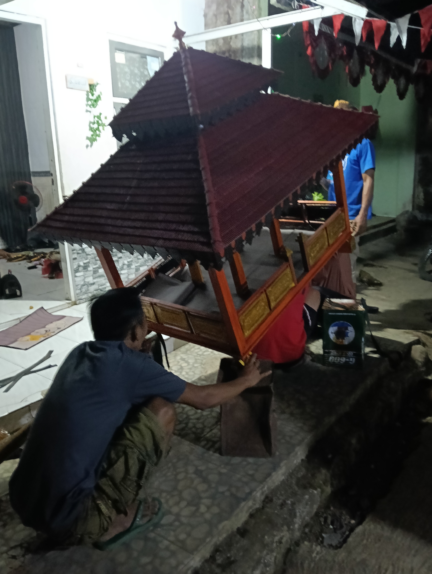 In the city of Tasikmalaya, Indonesia, on a ground-level outdoor patio, two men are seen working on a decorative wooden gazebo. One man, wearing a blue t-shirt and green camouflage pants, is crouched down, appearing to adjust a part of the gazebo's base. The second man, in a blue t-shirt, is standing behind the gazebo, partially obscured. The gazebo itself is ornate, with a dark red, multi-tiered roof and a wooden frame adorned with golden decorative elements. In the background, a light-colored wall with a window and some hanging greenery is visible, along with a fan and scattered items on the floor. Further back, red and white triangular flags are strung across a white structure, and a person wearing a red shirt is positioned behind the gazebo, possibly assisting. A green cylindrical object with a label showing an image and the text "609-9" is also present near the base of the gazebo. The ground is a mix of textured paving stones and rougher, darker surfaces.