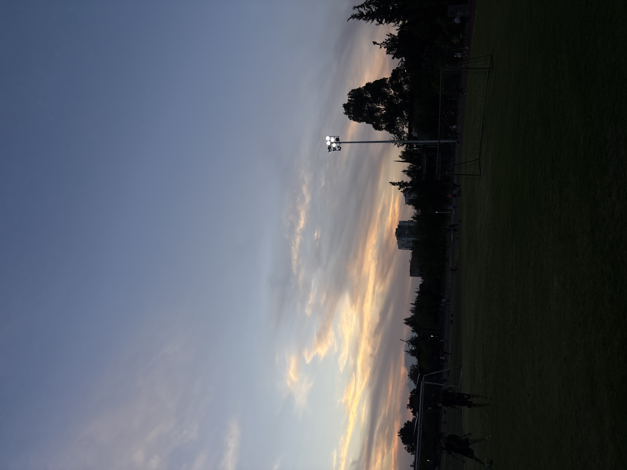 The image depicts a twilight scene at a football (soccer) field. The sky, occupying the upper and left portions of the frame, transitions from a deep blue at the top left to warm hues of orange, pink, and yellow in the elongated clouds stretching across the right side, indicating dusk or late evening.
A dark green grass field, likely a football pitch, covers the bottom and right sections. A tall light pole, equipped with multiple bright floodlights, stands prominently, illuminating the field against the fading natural light. Two football goals are visible on the field; one is larger and closer to the foreground, while a smaller one appears further back. The perimeter of the field is lined with the dark silhouettes of trees, and indistinct building outlines can be seen in the distant background on the right.
Several people are discernible on the field, appearing as dark silhouettes against the dim light. They seem to be engaged in a game, practice, or training session, with at least two figures clearly visible near the closer goal. The combination of artificial stadium lights and the colorful twilight sky creates a dynamic atmosphere. There is no visible text in the image. The scene suggests an active sporting event or practice taking place in the evening at an outdoor venue, potentially in Puebla, Mexico.
