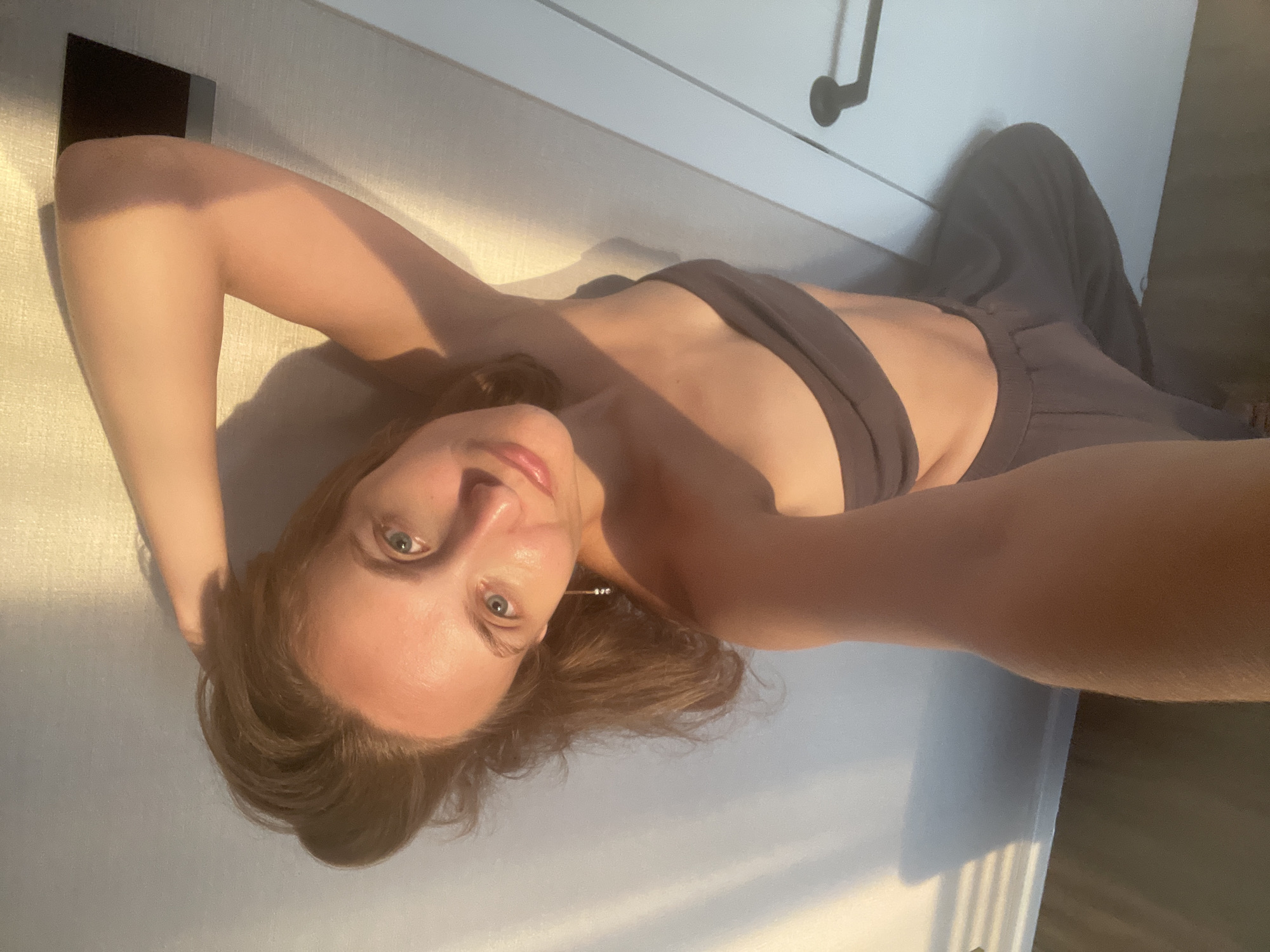A young woman with long, light brown hair and blue eyes is lying on her side, facing the camera. She is wearing a dark gray sports bra and dark gray sweatpants. Her left arm is bent, with her hand resting on her head. Her right arm is extended. The lighting suggests it is either sunrise or sunset, with long shadows cast across the scene. The background appears to be a room with light-colored walls and a white cabinet with black handles. A dark rectangular object is embedded in the wall to the left of the woman. The woman's expression is relaxed and serene.