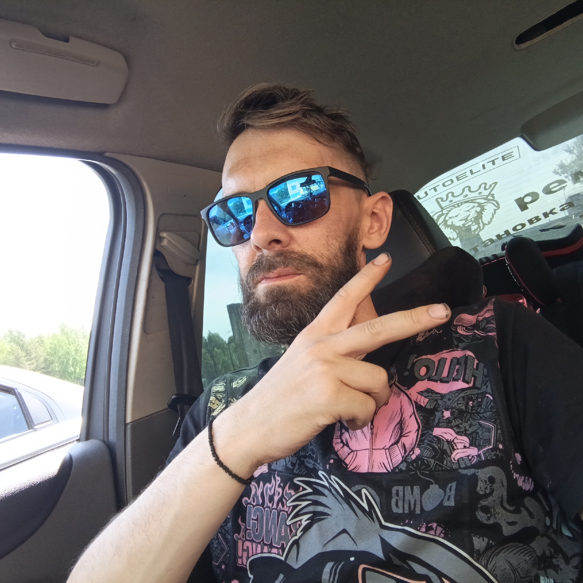 A man with a beard and sunglasses is sitting in a car, making a peace sign with his right hand. He is wearing a black t-shirt with colorful graphics and a black beaded bracelet on his left wrist. The interior of the car is visible, including the seat, seatbelt, and window. Through the window, trees and a car are visible. A sticker is on the rear window with the word "AUTOELITE" visible. The reflection in the sunglasses appears to show a building. The location is Markovskii, Russia.
