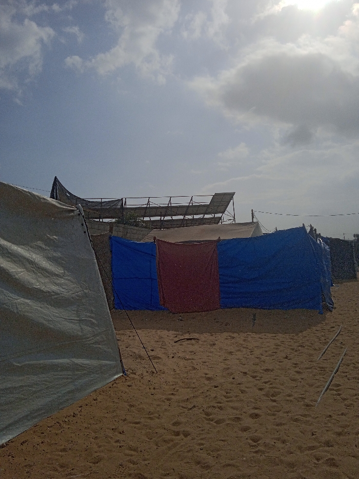 The image depicts a makeshift encampment or temporary settlement on sandy ground under a partially cloudy sky in Khan Yunis, Palestinian Territory. In the foreground, the lower left quadrant shows a portion of a light grey or beige tarp tent, taut and secured, with a dark line extending towards the ground. Directly ahead in the midground, a temporary barrier or wall is constructed from three vertical tarps: two blue tarps flank a central reddish-brown tarp. These tarps are strung across an unseen framework. In the background, behind the tarp wall, a more permanent structure is visible, featuring a frame that supports dark mesh netting and sloped panels, possibly solar arrays. Concrete blocks are also evident as part of this background structure. The sandy ground extends across the entire lower half of the frame, showing minor undulations and sparse debris, including two light-colored rods on the right side. No individuals or specific activities are visible within the scene.