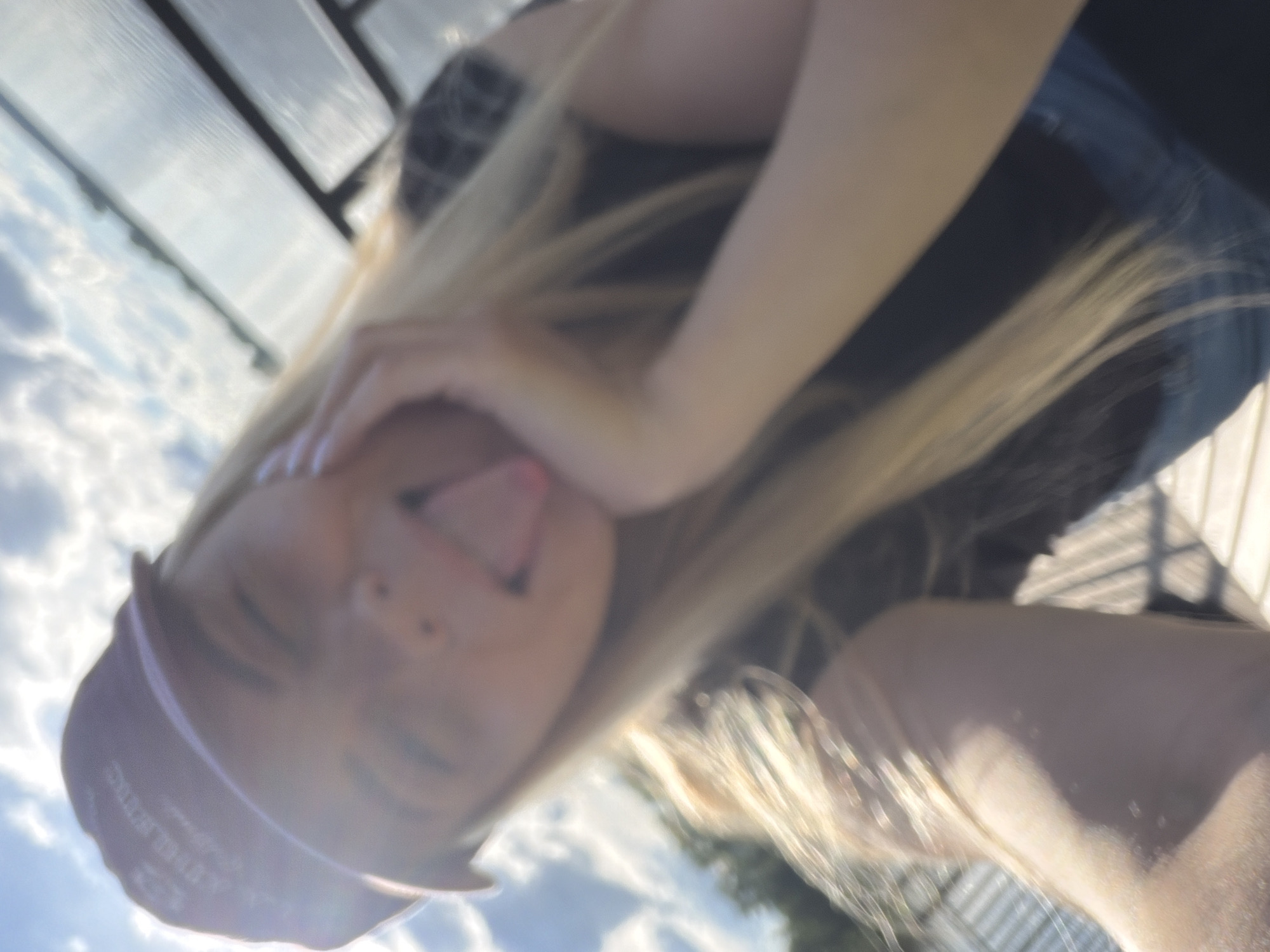 A blonde-haired individual with a purple headscarf, wearing what appears to be a black tank top and jeans, sticks out their tongue in a selfie taken from a low angle. The sky with clouds is visible in the background. The location is unspecified and cannot be determined as Votkinsk, Russia.