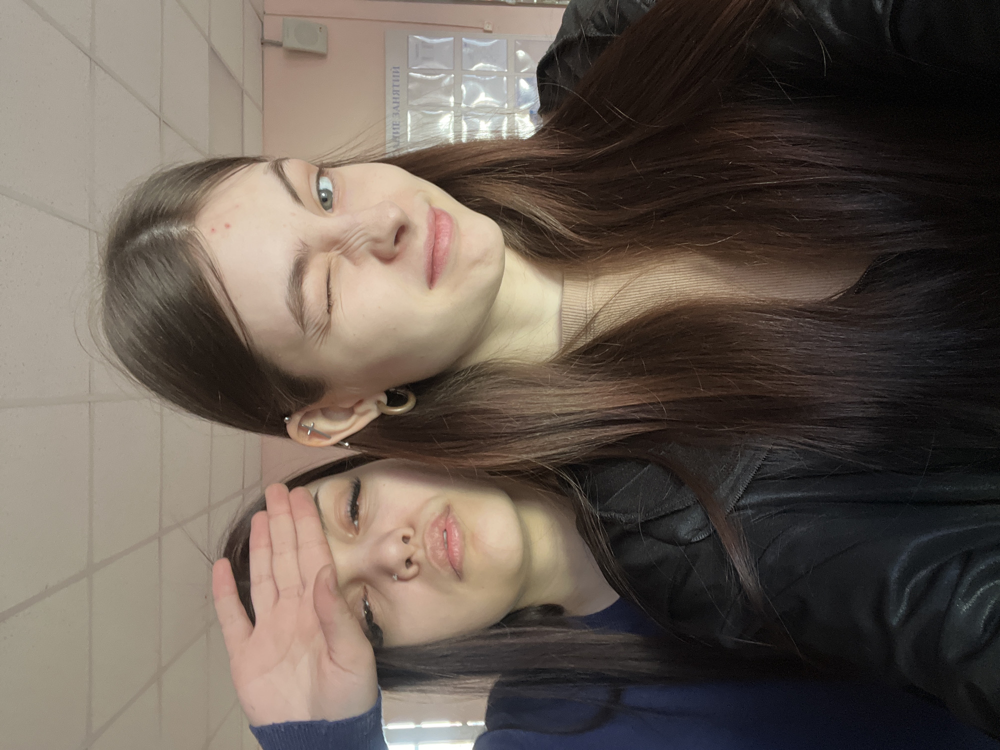 Two young women are posing for a photo indoors. The woman on the top is winking her left eye. Both women have long brown hair and are wearing dark-colored jackets. The background consists of a tiled wall and what appears to be a bulletin board with translucent paper covers. The location is likely Russia, as indicated by the text on the bulletin board which is written in Russian. Given the interior setting and the lack of specific landmarks, determining if the image was taken in Saint Petersburg is not possible without additional information.