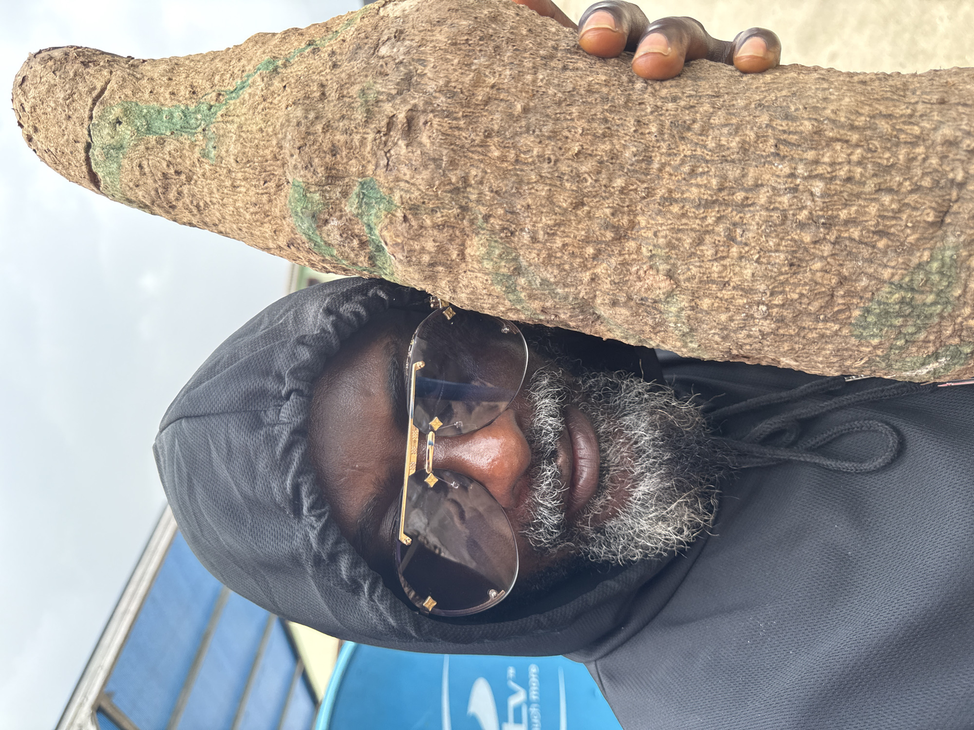 In the city of Ado, Nigeria, a close-up shot captures a man in a black hooded sweatshirt and large, gold-accented sunglasses. He holds a large, gnarled root or piece of wood in his right hand, with his fingers visible against the textured surface. The root has green markings on it. A blue object with white lettering is visible in the background to the left. The sky is overcast. The man has a graying beard and mustache.