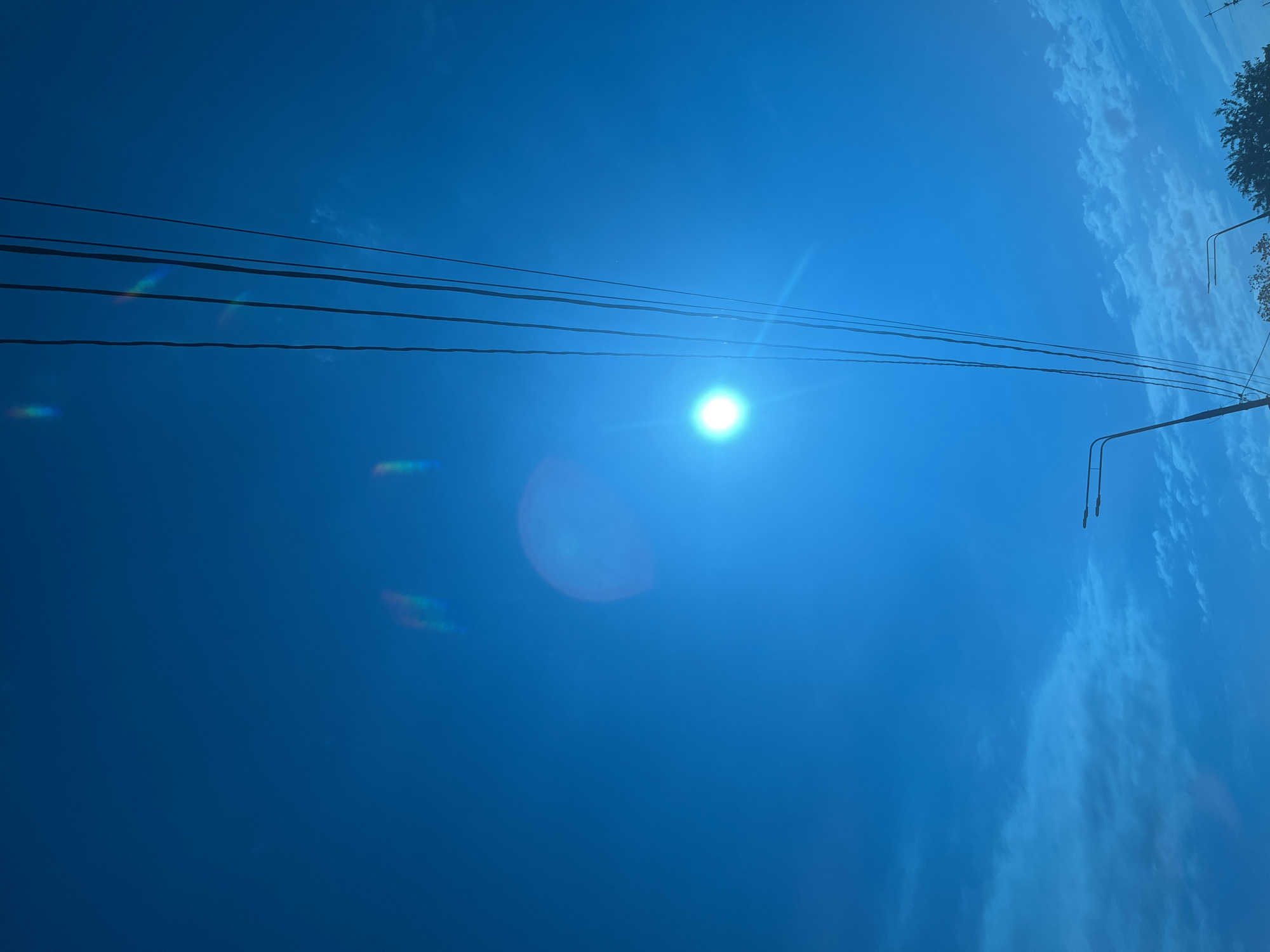 The image shows a bright blue sky with the sun shining intensely. Several power lines run across the top left of the frame to the center. To the right, there is a utility pole with two curved extensions. Light cloud formations are visible in the top right corner. The image is taken from a low angle, looking up. There are no people visible. It is impossible to determine the location of the image in the city of Krasnodar, Russia, or any other specific location from the information given.