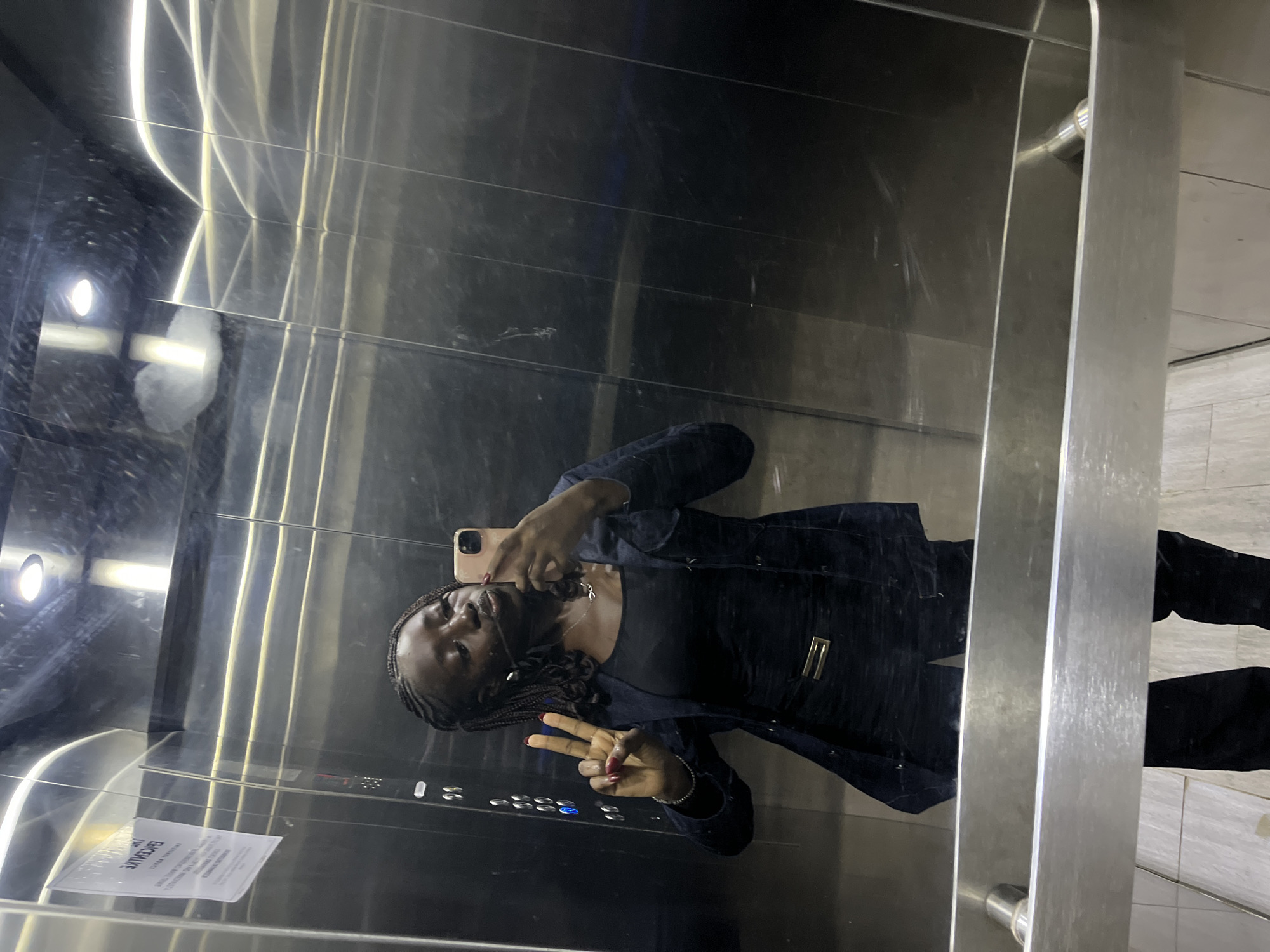 A person is taking a selfie in an elevator with shiny metal walls. The individual is wearing a dark-colored outfit and holding a phone up to the mirror. They are giving a peace sign with their hand. There's a paper sign posted on the elevator wall, and the elevator buttons are visible. Based on the prompt information, the location is Karum, Nigeria.
