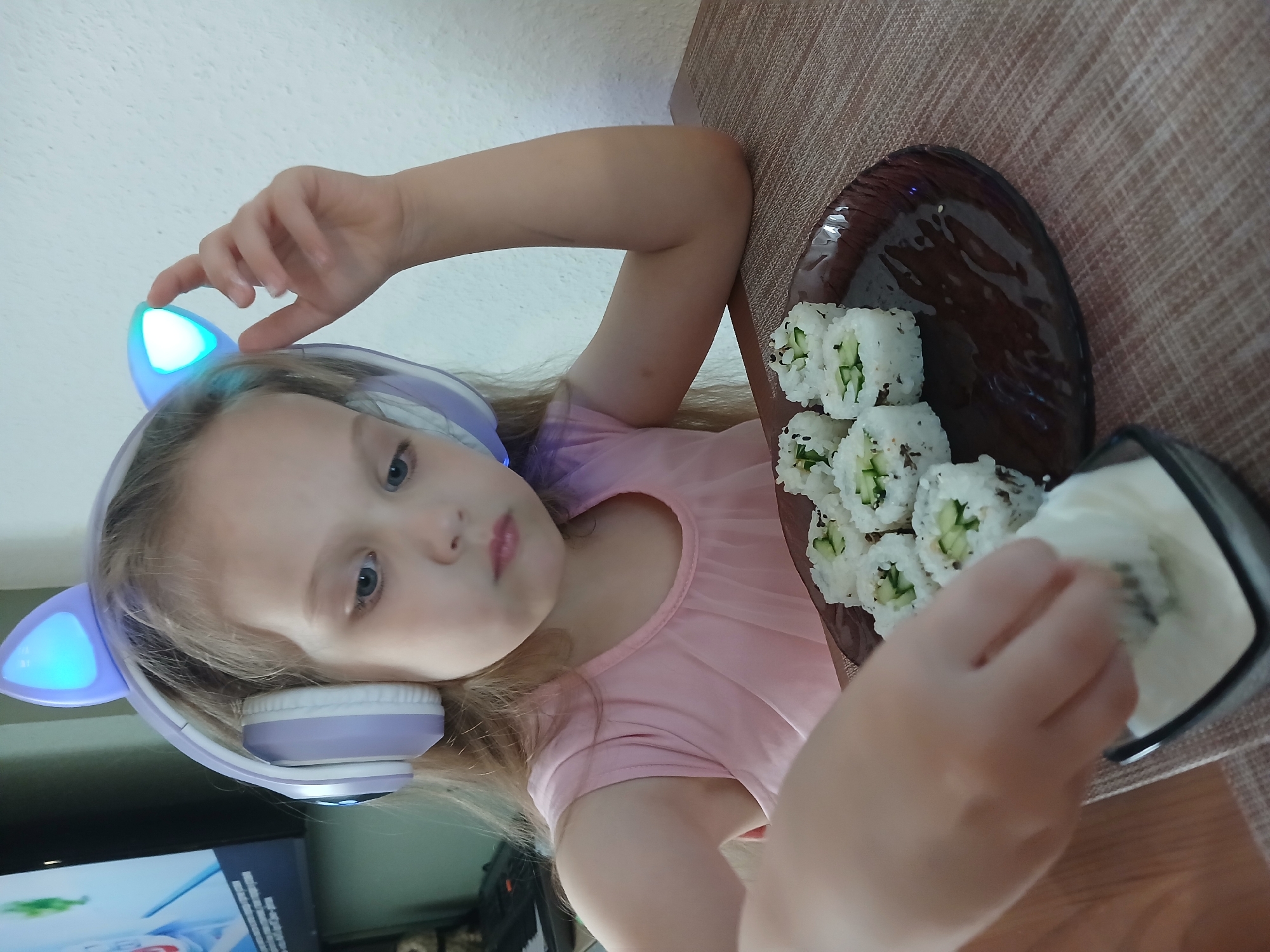 A young girl with blonde hair, wearing purple headphones with light-up cat ears, sits at a table and is about to dip a sushi roll into a white sauce. She is wearing a pink sleeveless dress. In front of her is a plate with several sushi rolls. The background shows a wall and a television. The scene is indoors. The image does not provide any information about its location in Yekaterinburg, Russia.