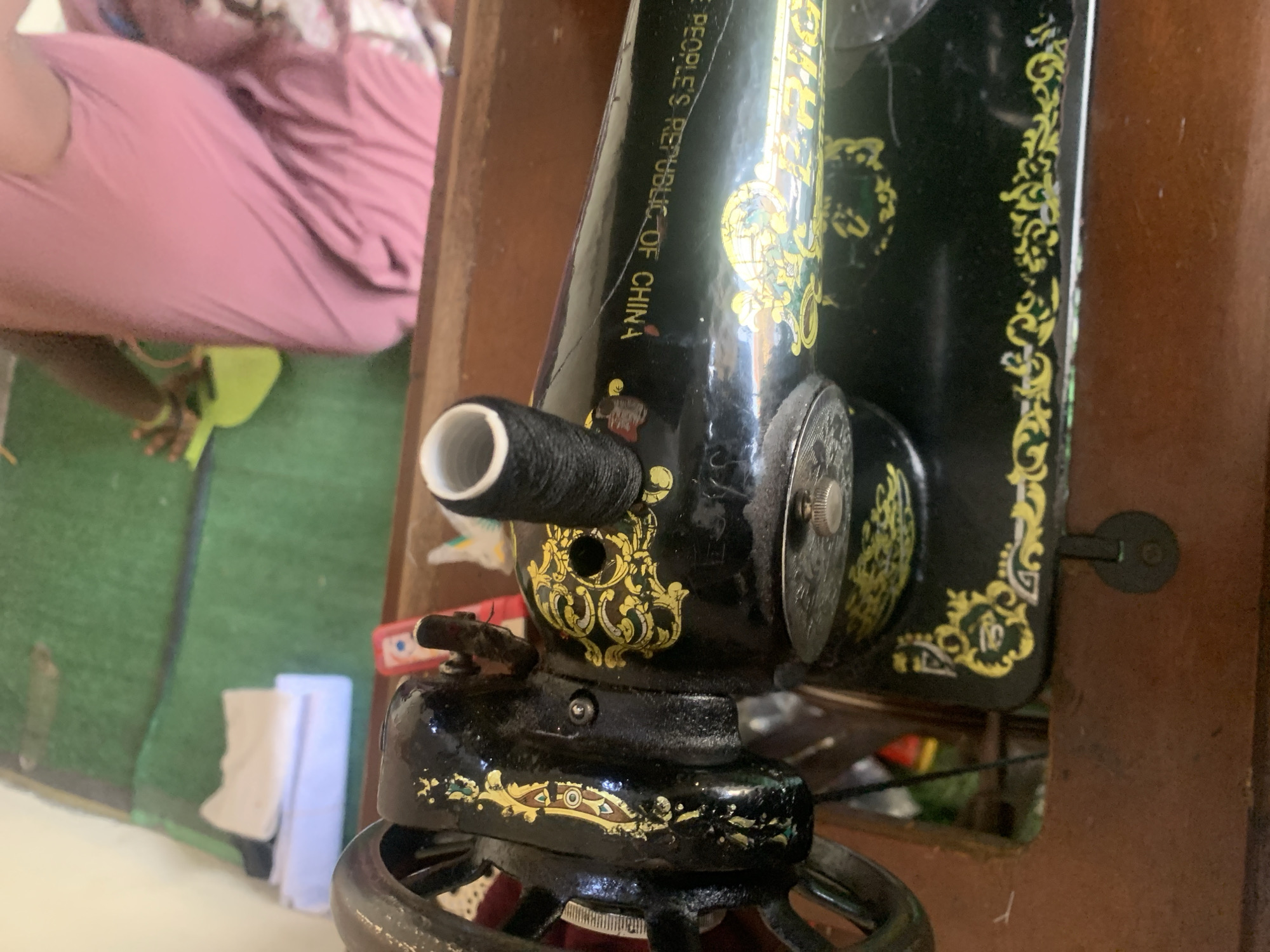 The image shows a close-up of a vintage black sewing machine, decorated with gold ornamentation. A black spool of thread is mounted on a spindle near the top of the machine. The sewing machine is situated on a wooden surface, possibly a sewing table or cabinet. In the background, a person wearing a pink garment is partially visible, and a green mat is also discernible. The location is identified as Jauro Yinu, Nigeria.
